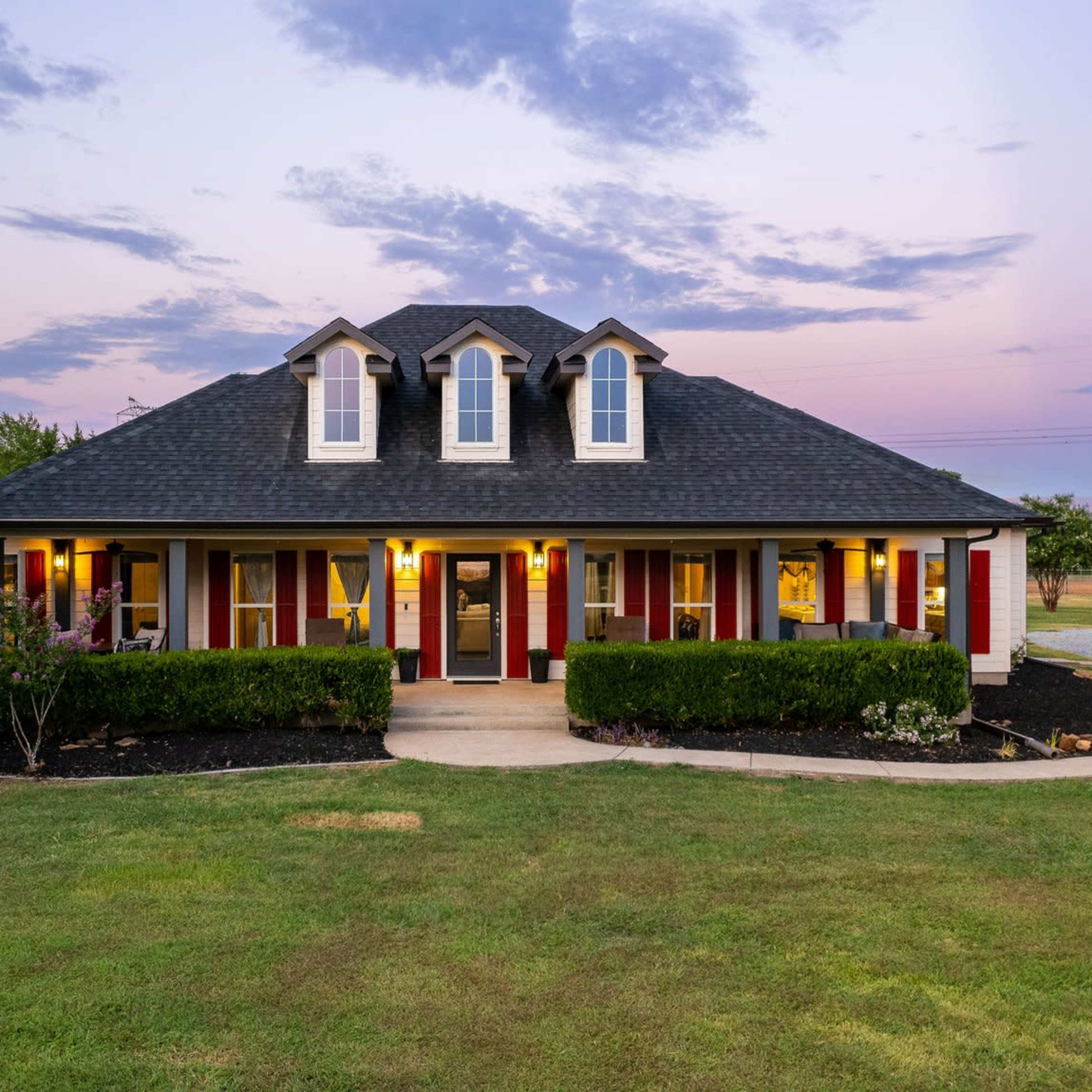 A two-story house with a dark roof and red shutters is surrounded by manicured lawns and shrubs under a twilight sky.