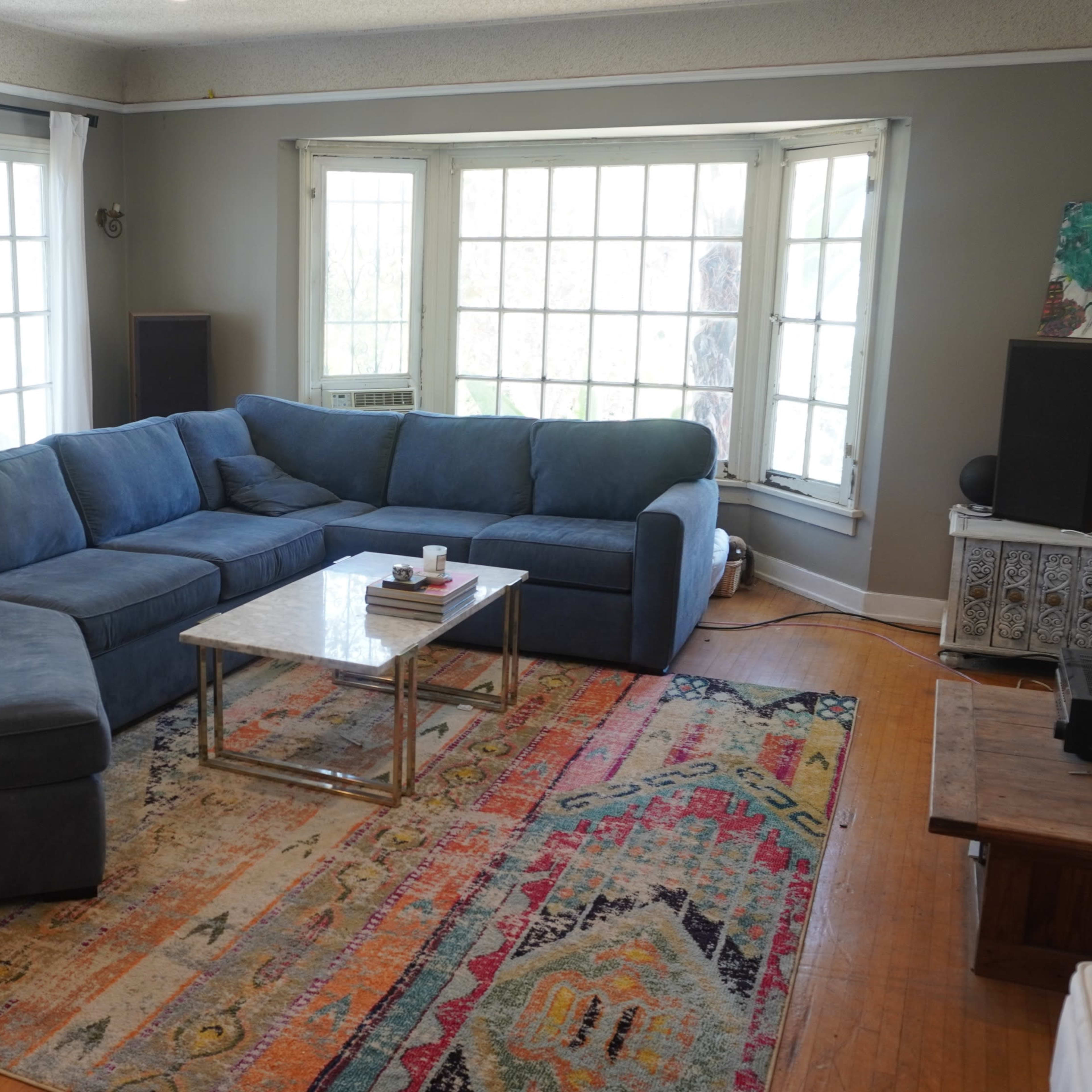 A spacious living room features a large blue sectional sofa, a glass coffee table, and colorful area rugs, with bright natural light streaming in through multiple windows.