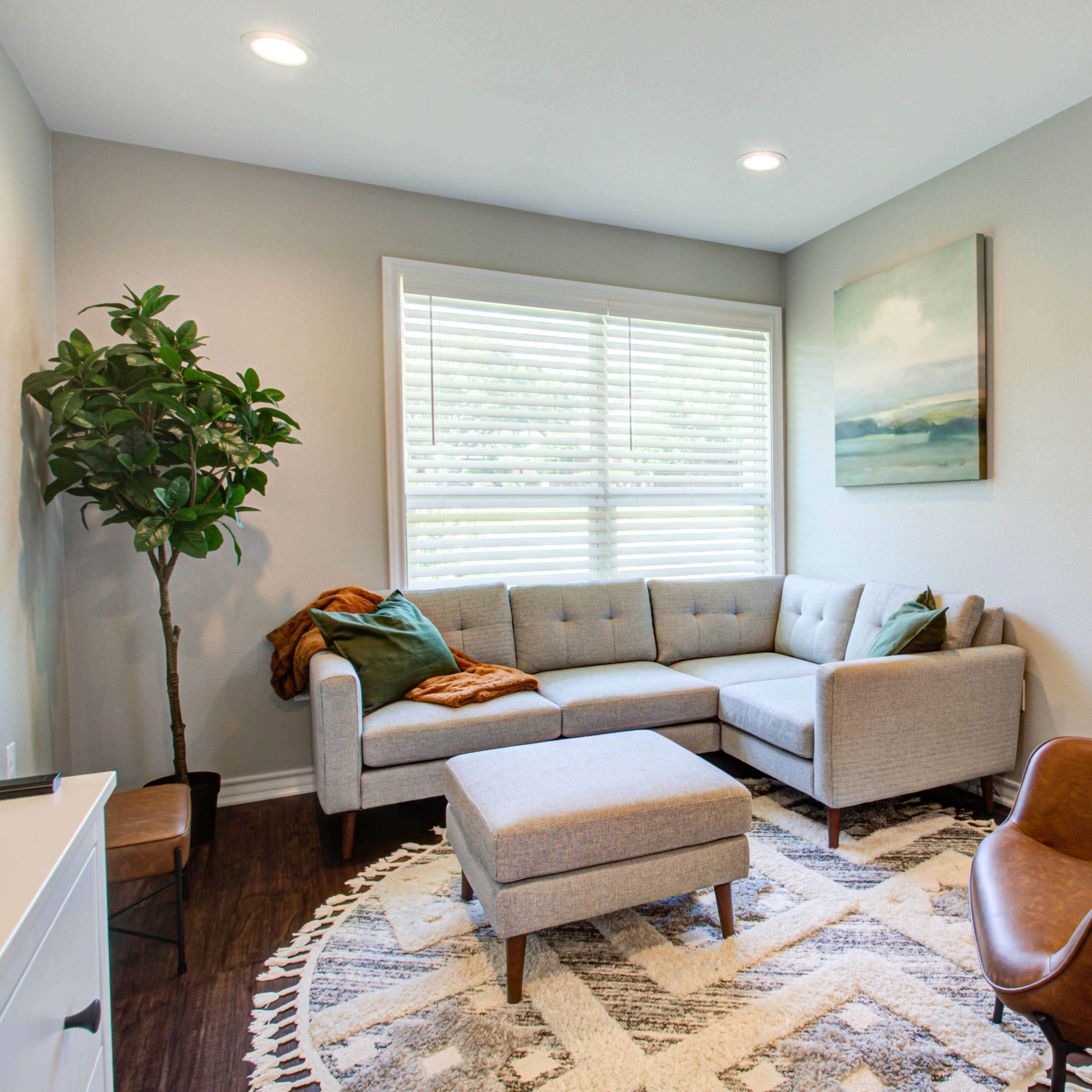 A modern living room features a grey sectional sofa, a brown leather armchair, a round patterned rug, and a large window with blinds.