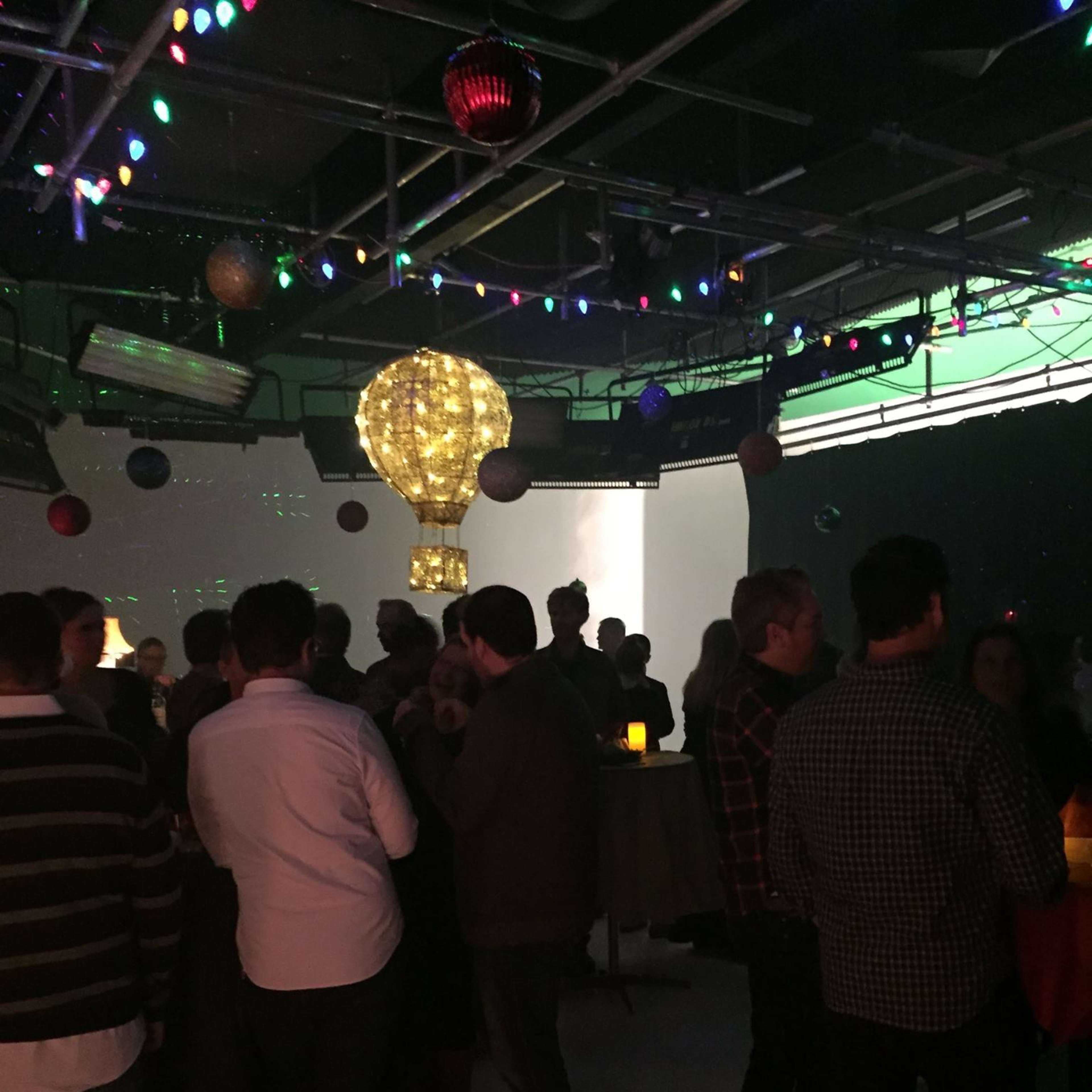 A lively gathering takes place indoors, with people mingling under colorful lights and a decorative hot air balloon hanging from the ceiling.