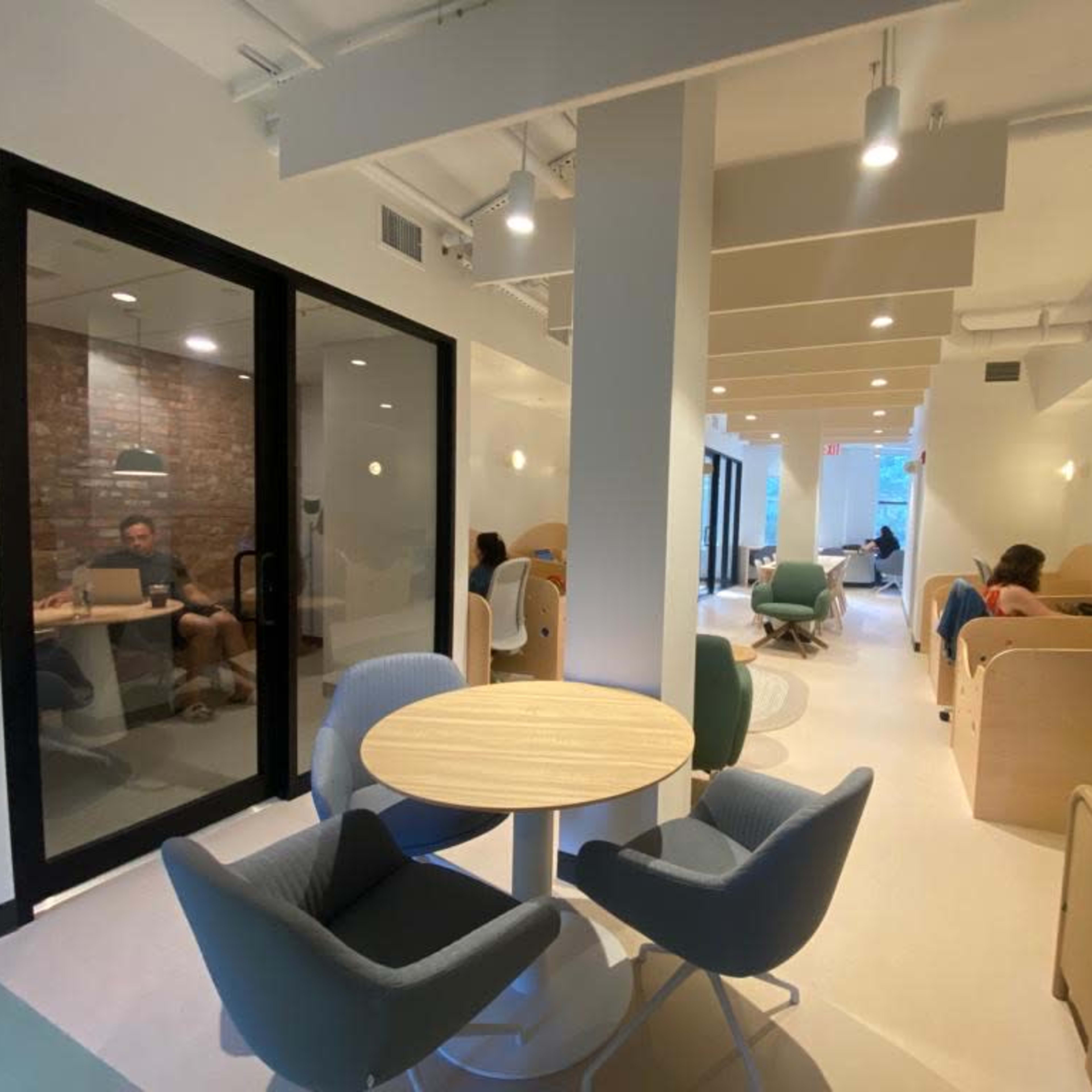 A modern coworking space with a mix of private meeting areas and open seating, featuring round tables and various seating arrangements.