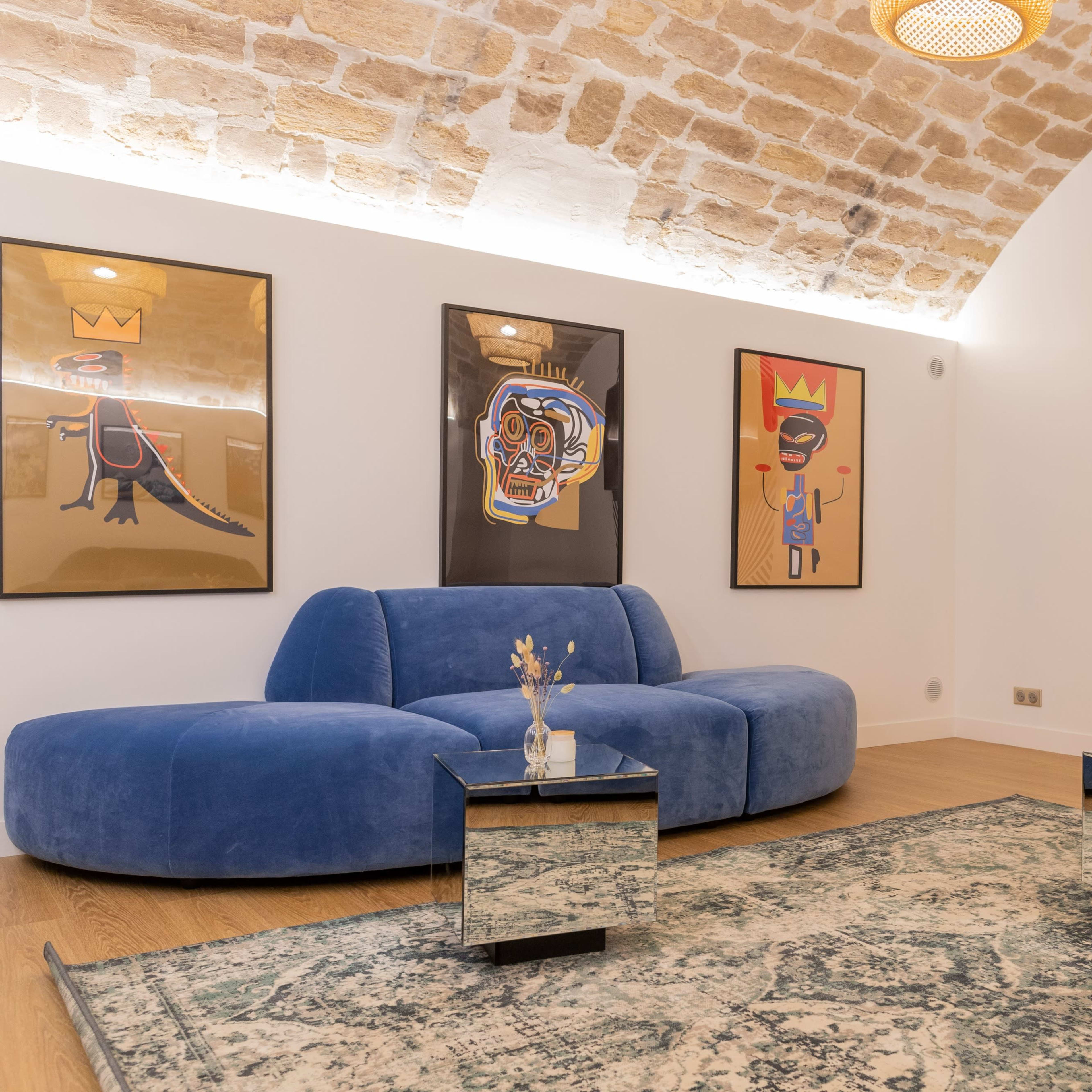 A modern living room featuring a blue curved sofa, two mirrored coffee tables, and colorful framed artwork on the walls, with a television mounted in the corner.
