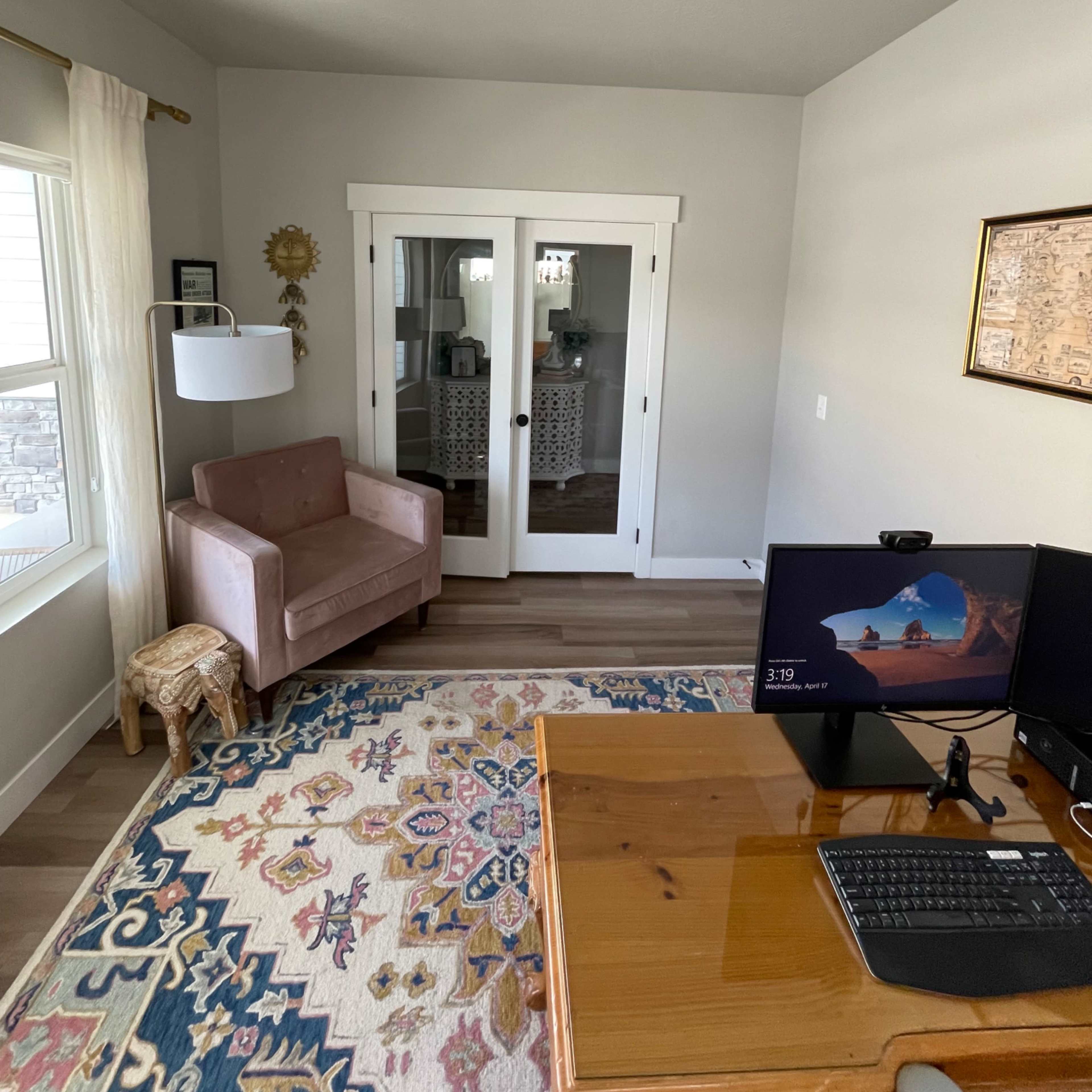 A small home office features a wooden desk with a monitor, a plush armchair, and a decorative rug, along with glass-paneled doors leading to another room.
