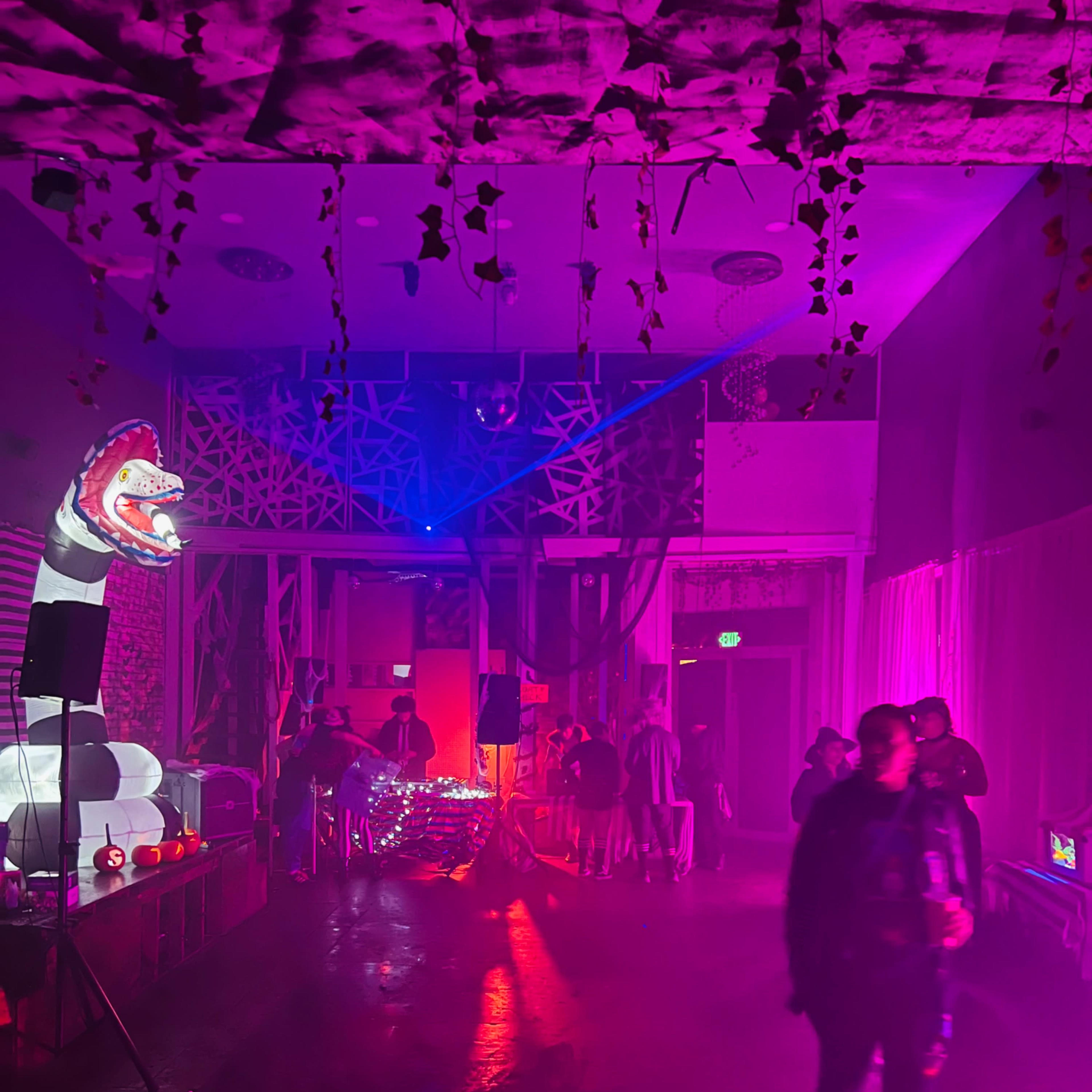 A vibrant indoor space features colorful lighting, a large dinosaur statue, and people mingling amidst music equipment and decorations.