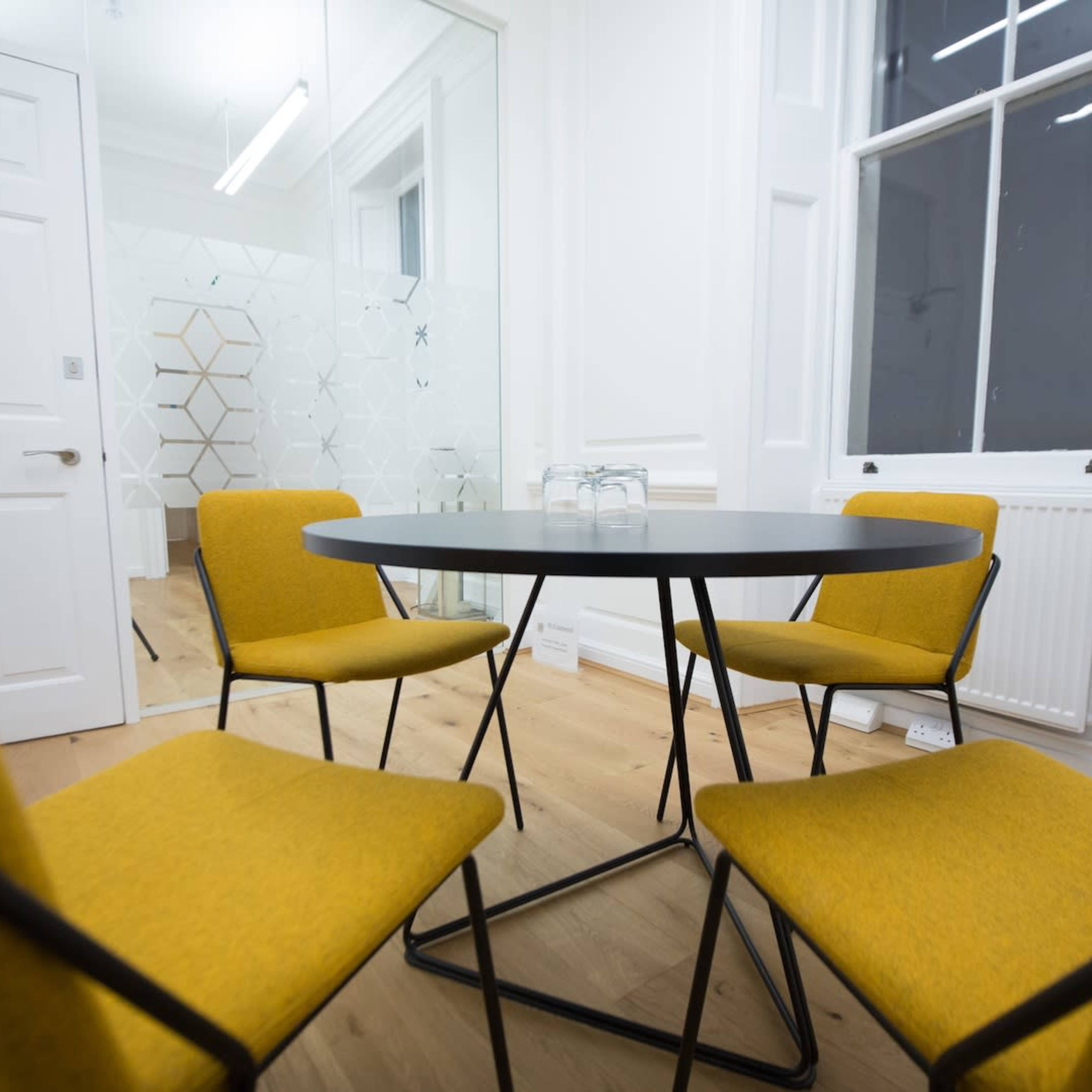 A round table is surrounded by four yellow chairs in a well-lit room with white walls and a large window.