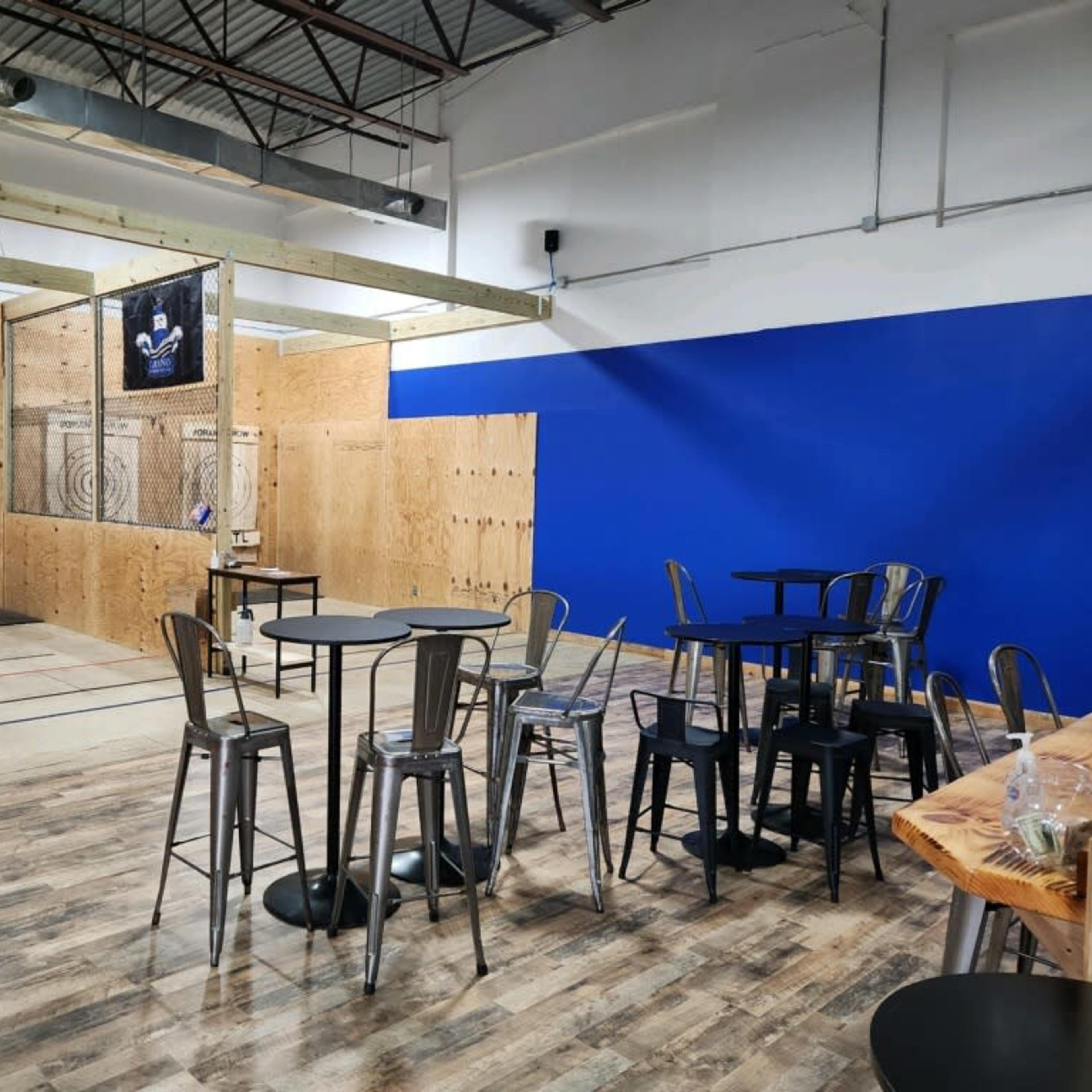 A spacious indoor venue featuring a wooden floor, multiple axe-throwing targets, and seating areas with metal tables and chairs.