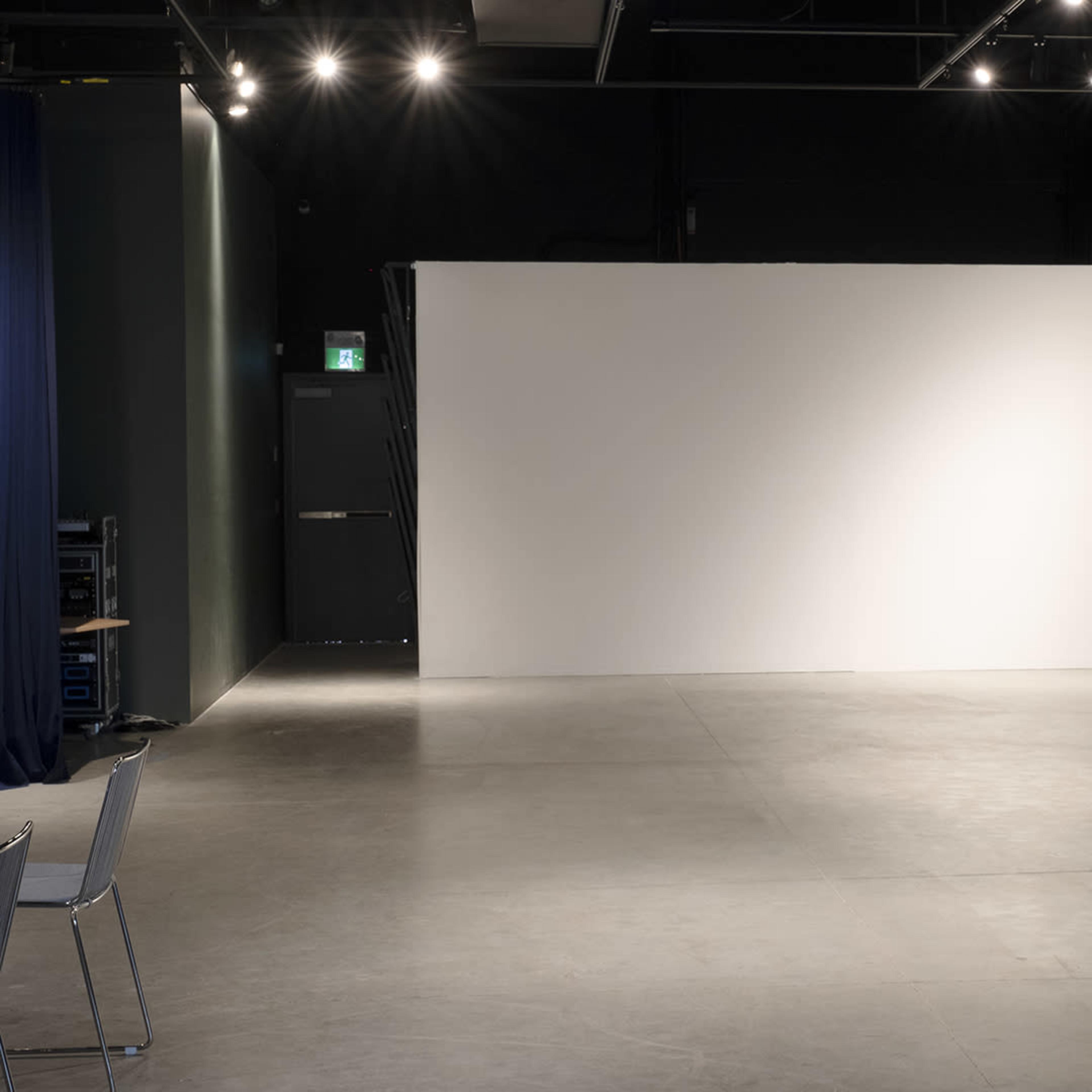 An empty performance space with a bare white wall and minimal furnishings.
