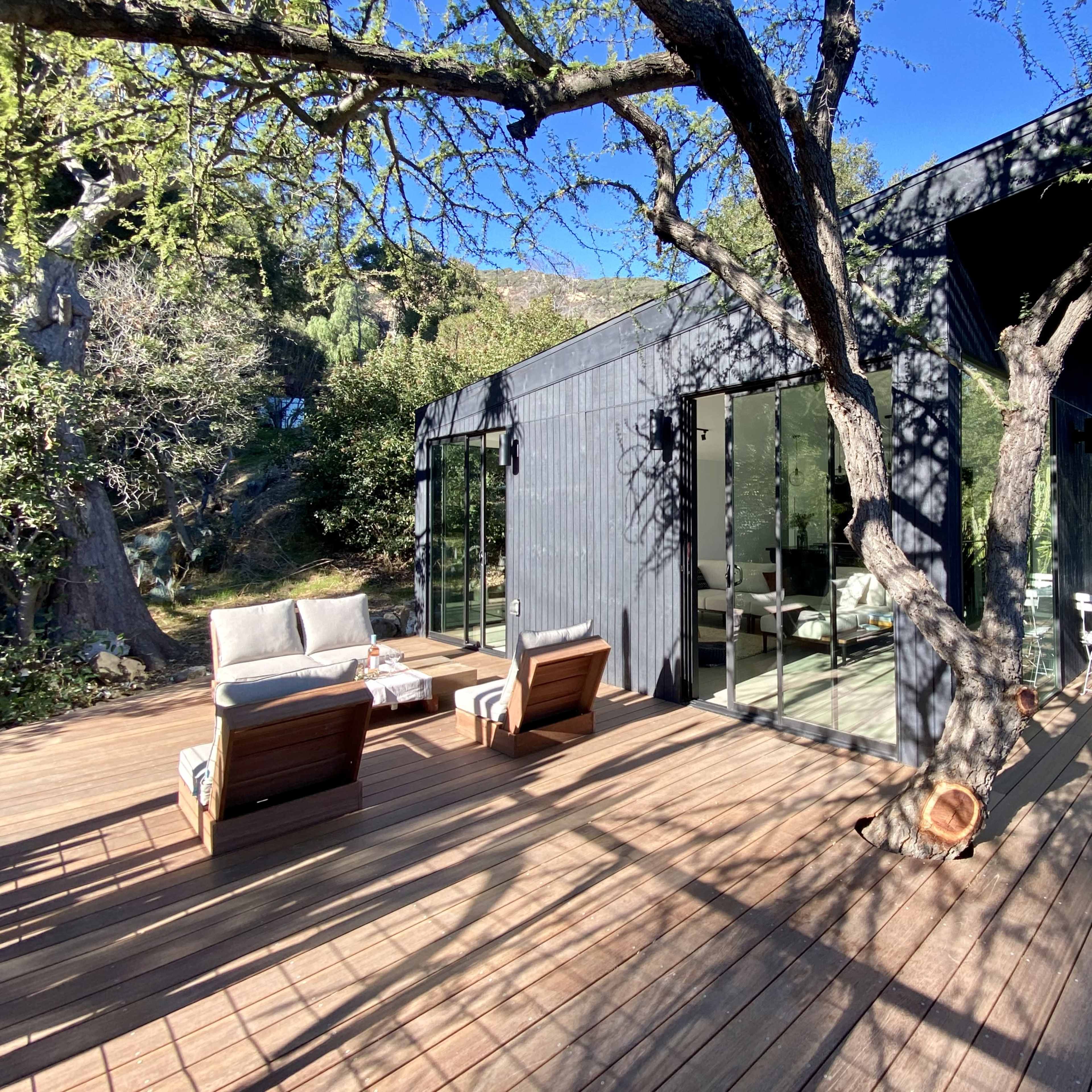 A modern black wooden house is surrounded by trees, featuring a spacious deck with seating arrangements.