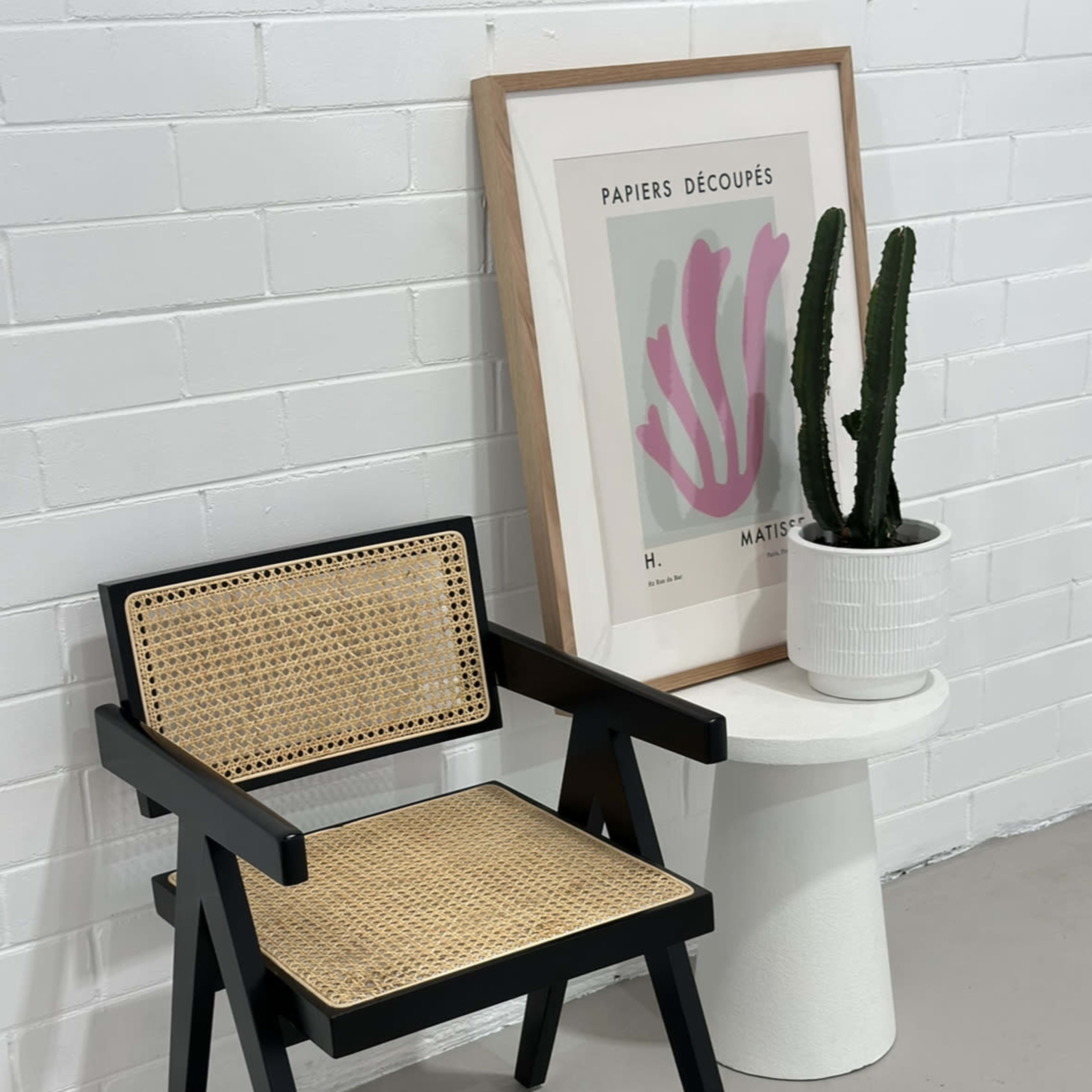 A black wooden chair with a woven seat is positioned beside a white abstract art print and a potted cactus on a cylindrical table.