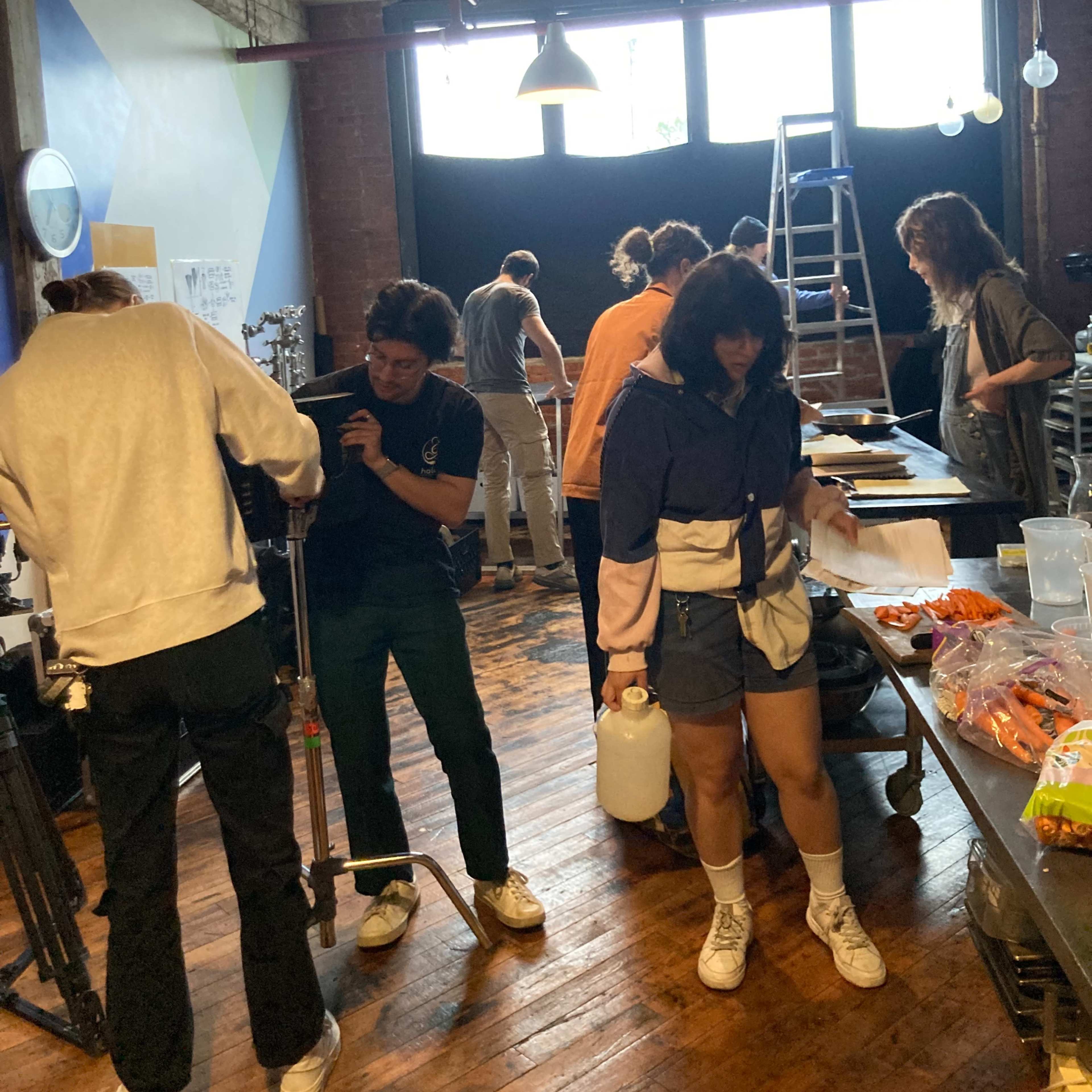 A group of people work on various tasks in a room with wooden floors and large windows, while some equipment and snacks are spread out on tables.