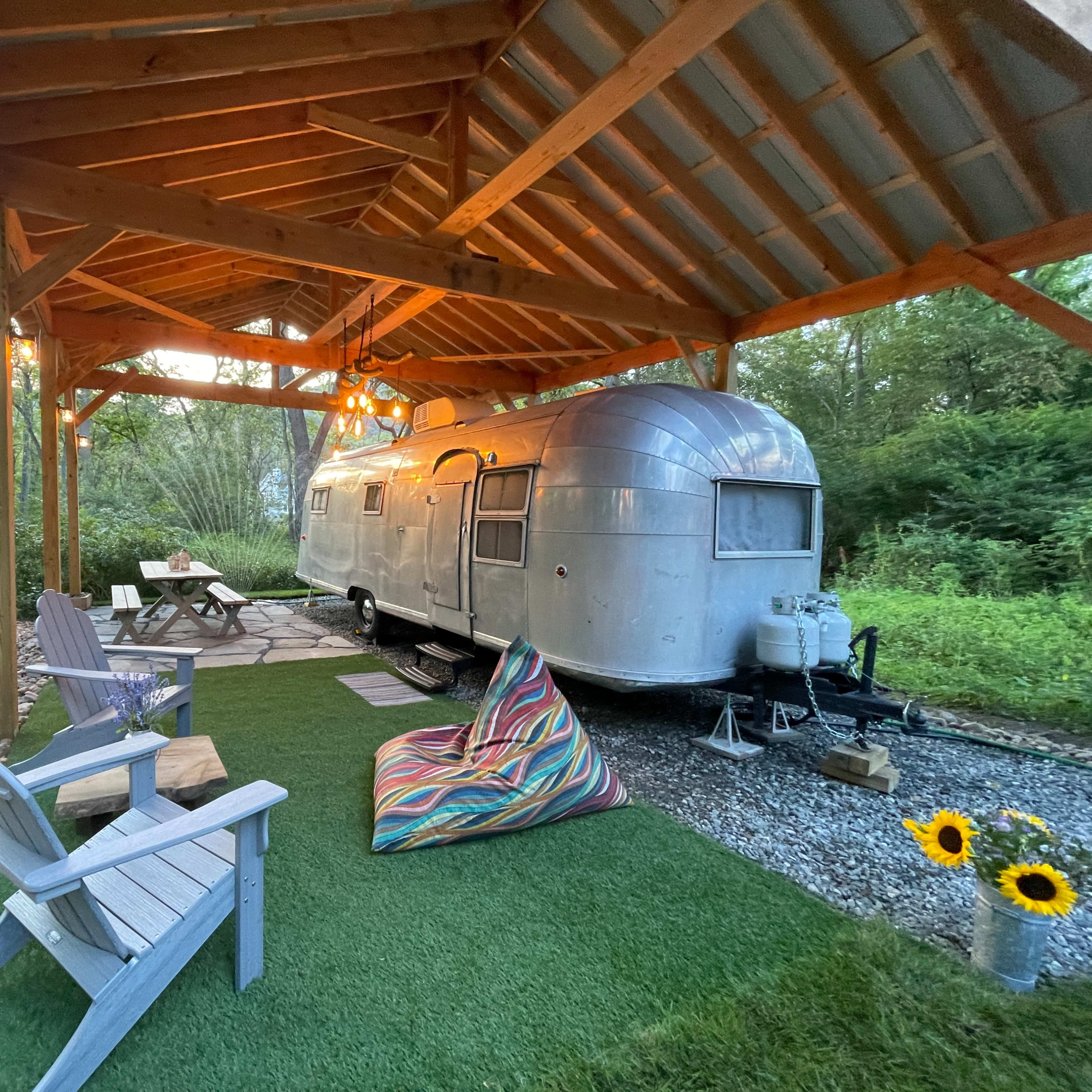 A silver trailer is parked under a wooden shelter with outdoor seating and a landscaped area featuring grass and decorative plants.