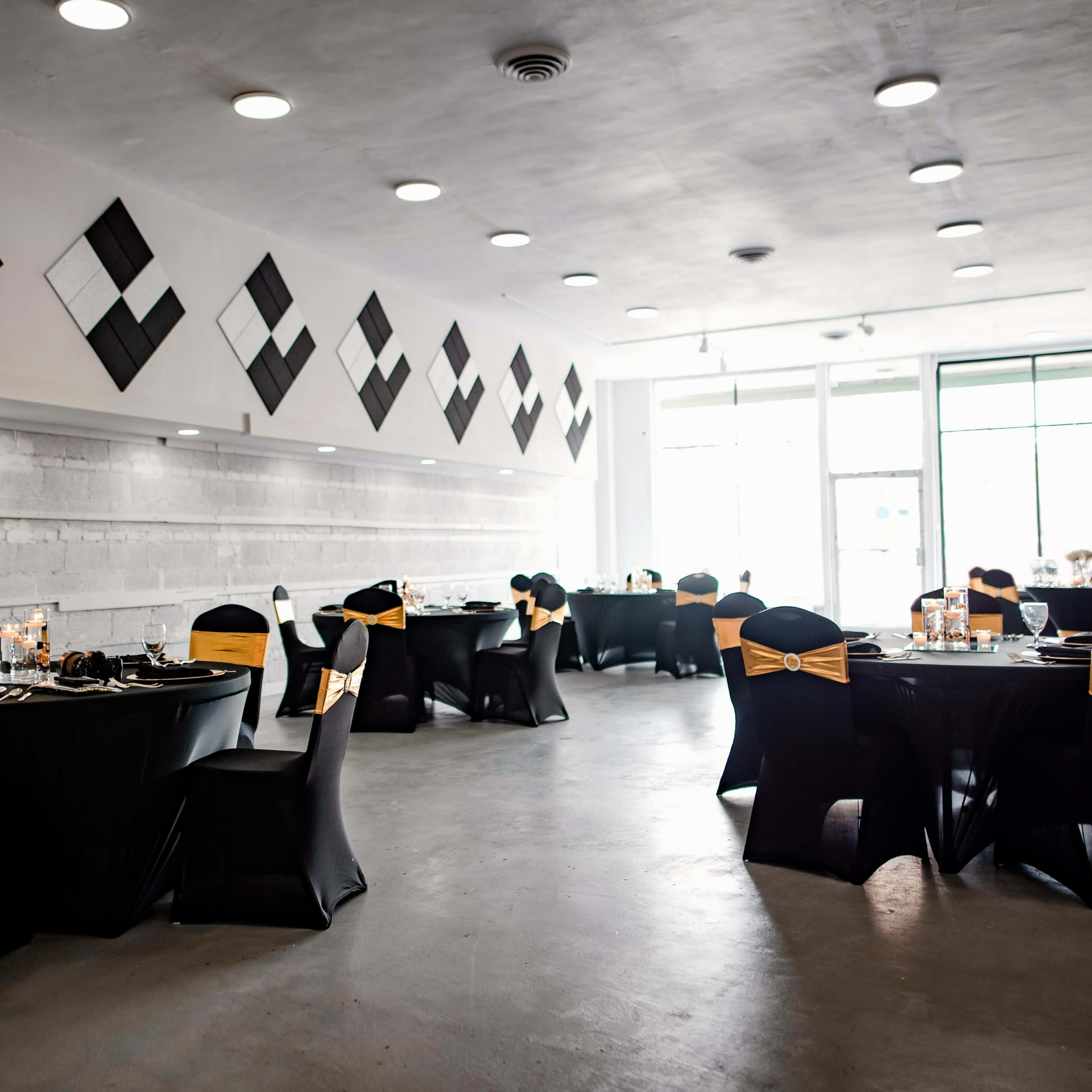 A modern event space set up with black tables and chairs adorned with gold sashes, and minimalistic decor on the walls.