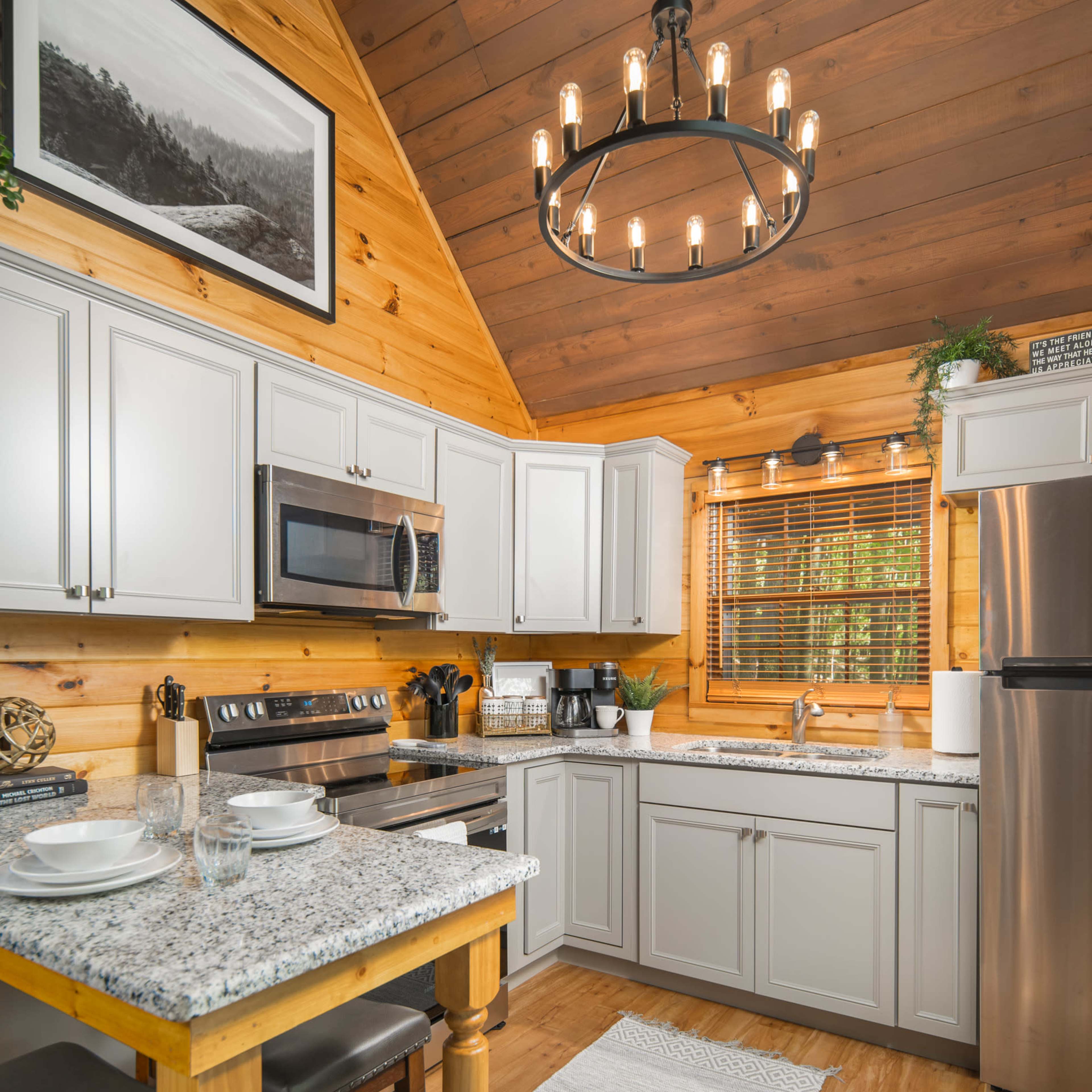 A modern kitchen with light gray cabinets, granite countertops, a stainless steel refrigerator, and wooden accents, all set under a chandelier in a cabin-style interior.