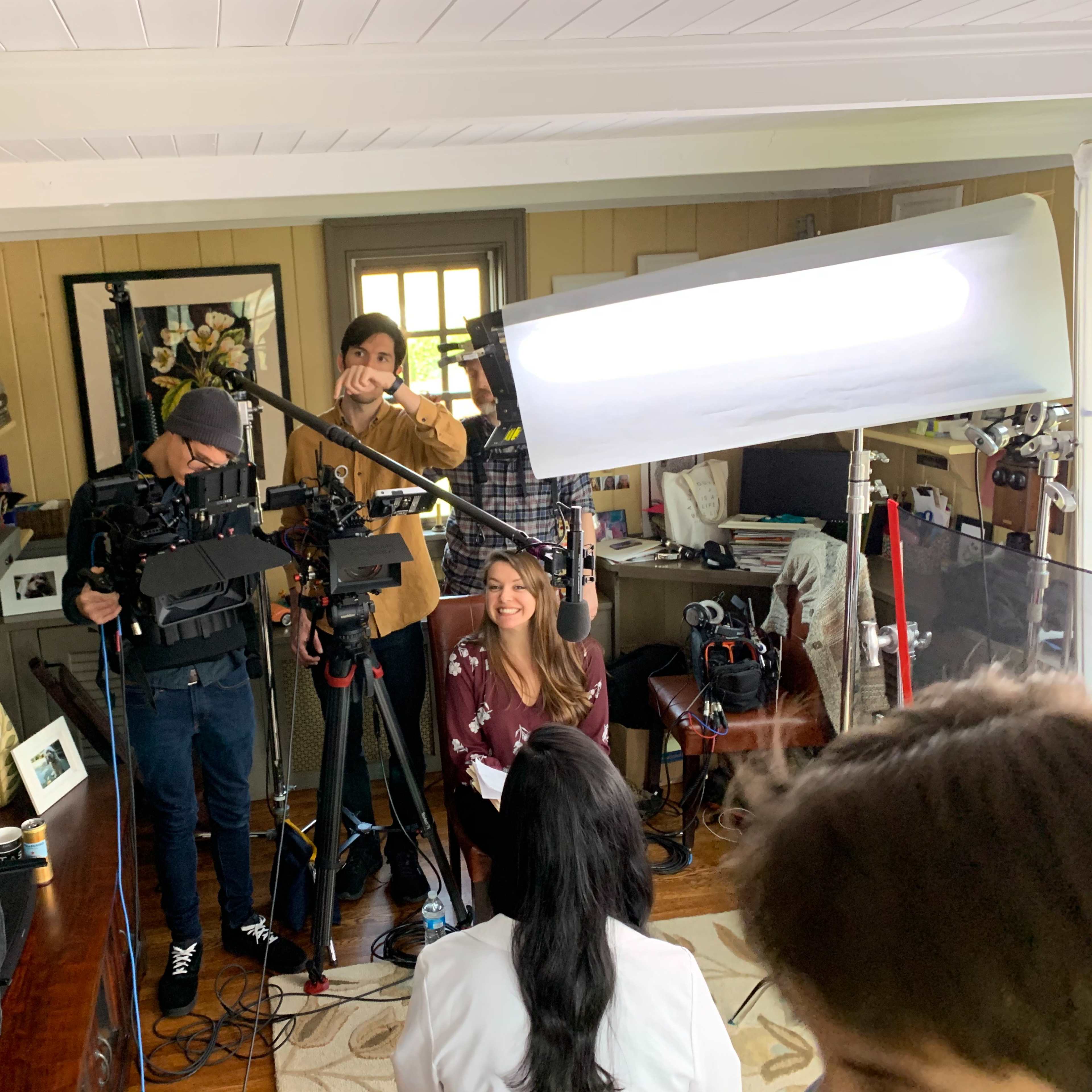 A video crew sets up equipment in a room for an interview with a woman seated in front of the camera.
