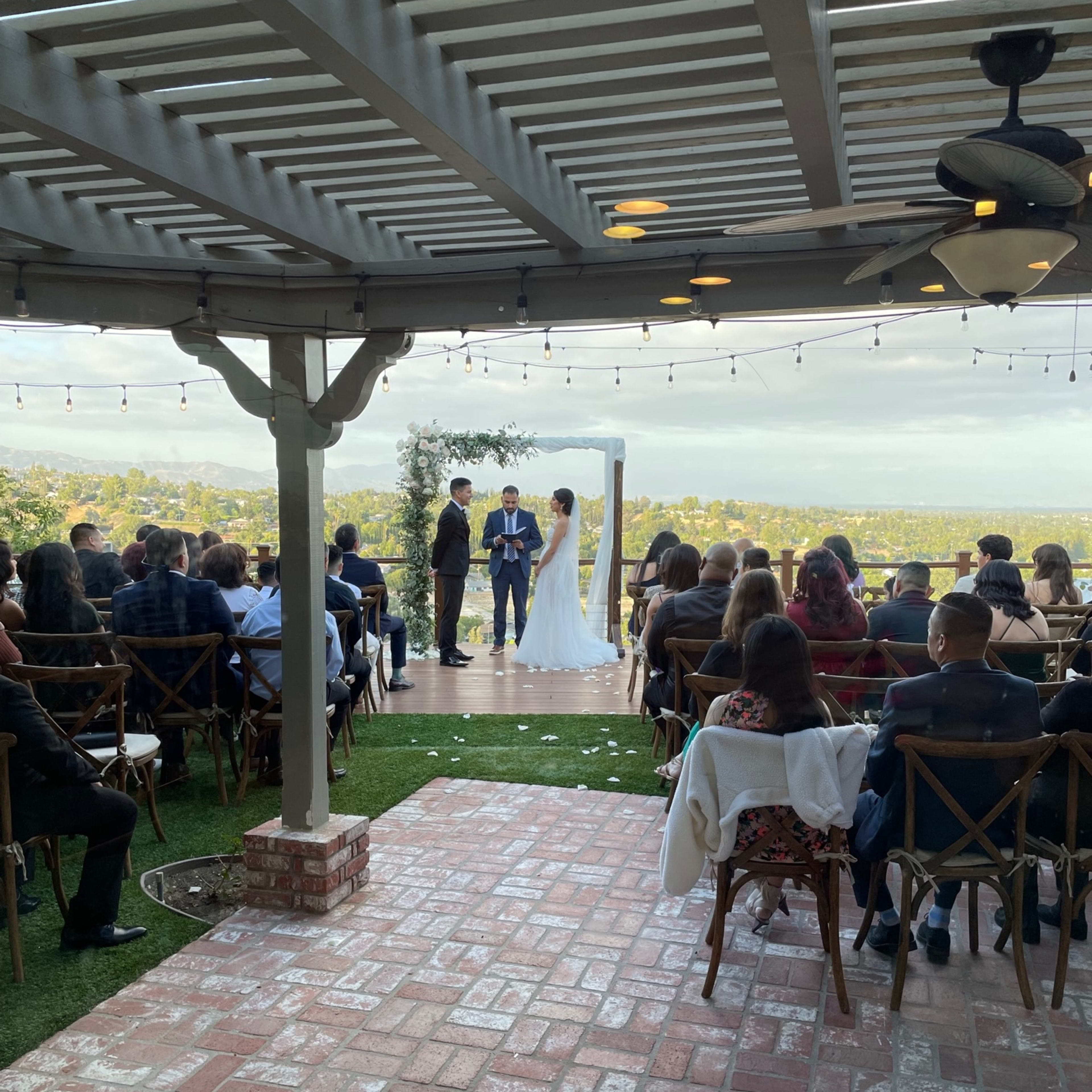 A couple stands under a canopy exchanging vows during their wedding ceremony, while guests are seated in rows on a patio overlooking a scenic landscape.