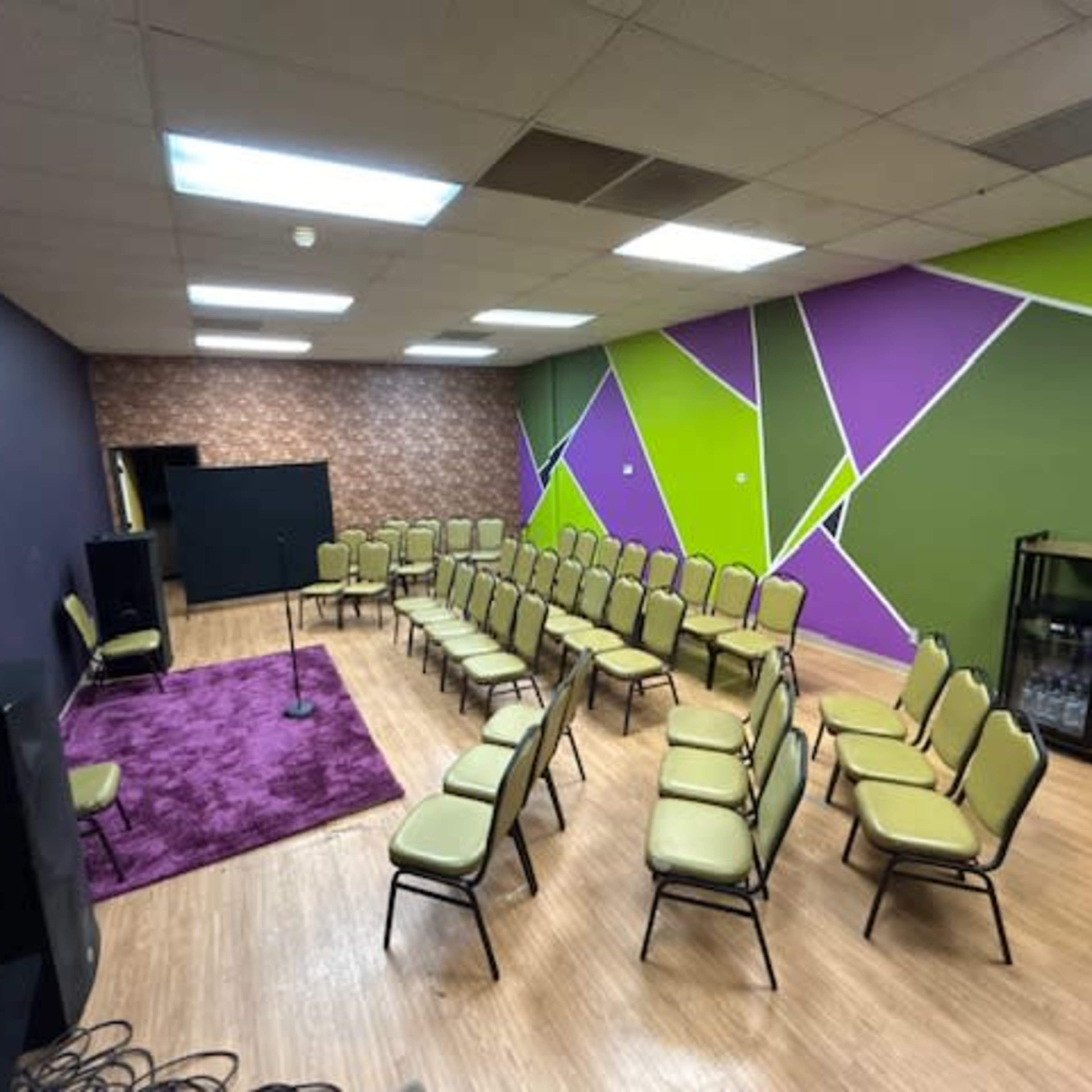 A spacious room arranged with rows of chairs facing a small stage, featuring colorful walls with green and purple geometric patterns.