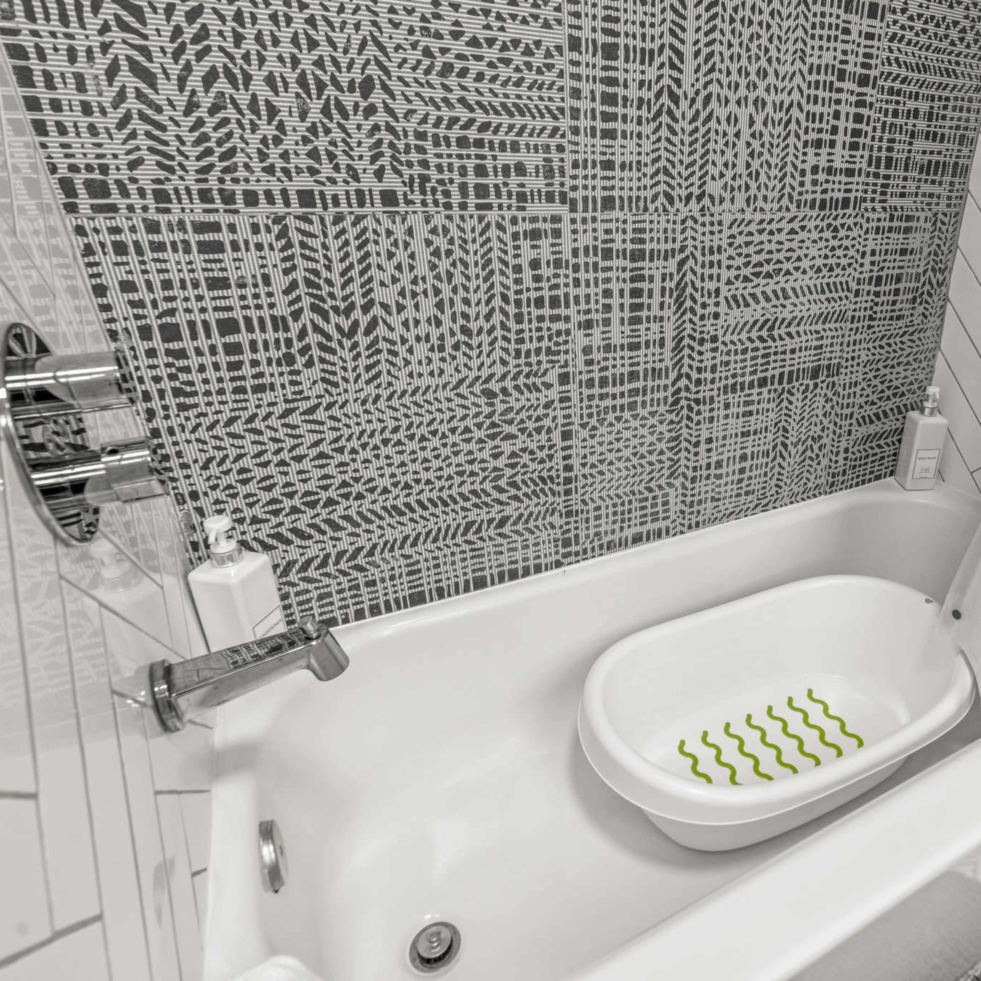 A white bathtub with a patterned mat inside, surrounded by tiled walls featuring a geometric design.
