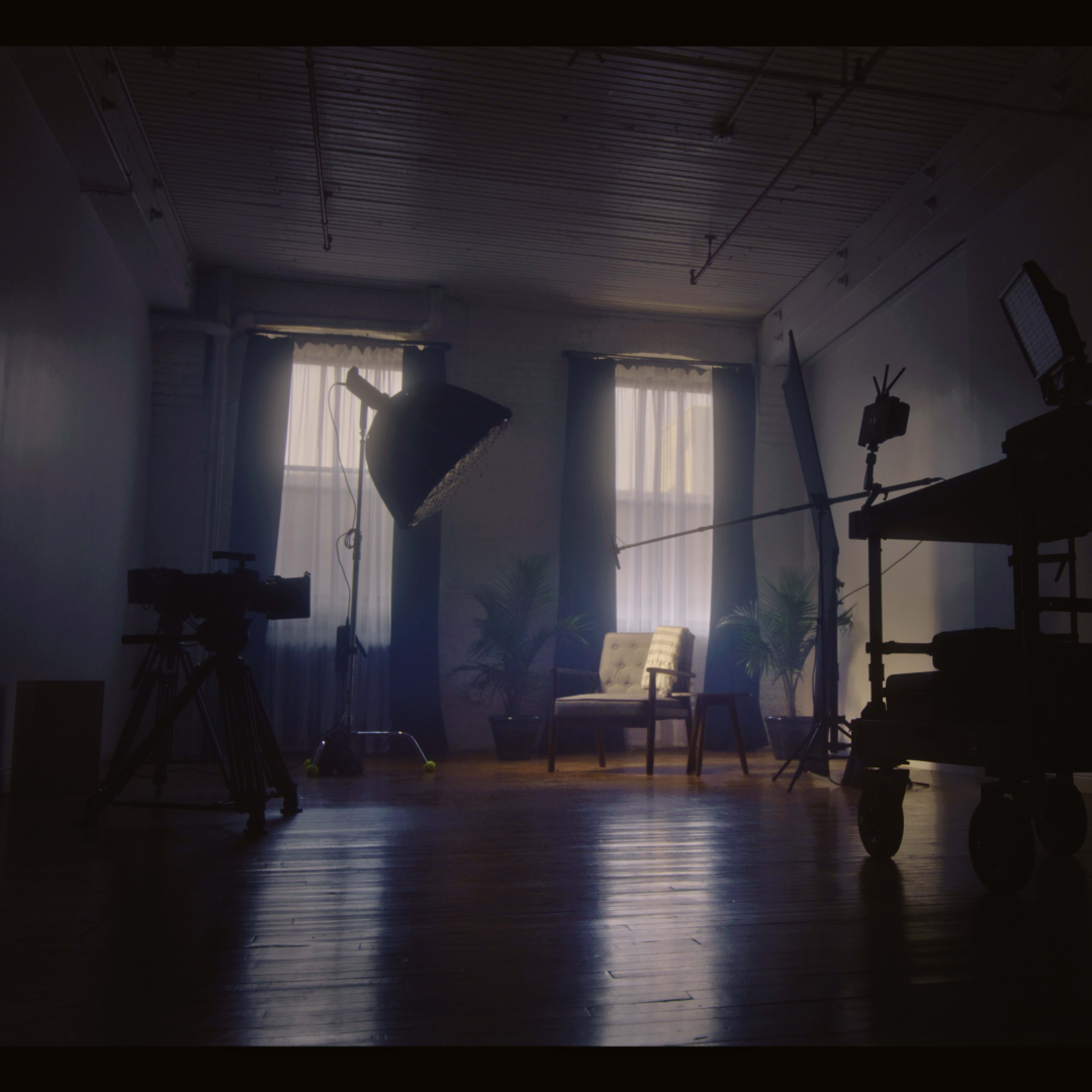 A dimly lit room features an empty chair illuminated by natural light and surrounded by studio equipment, including a camera and a light stand.