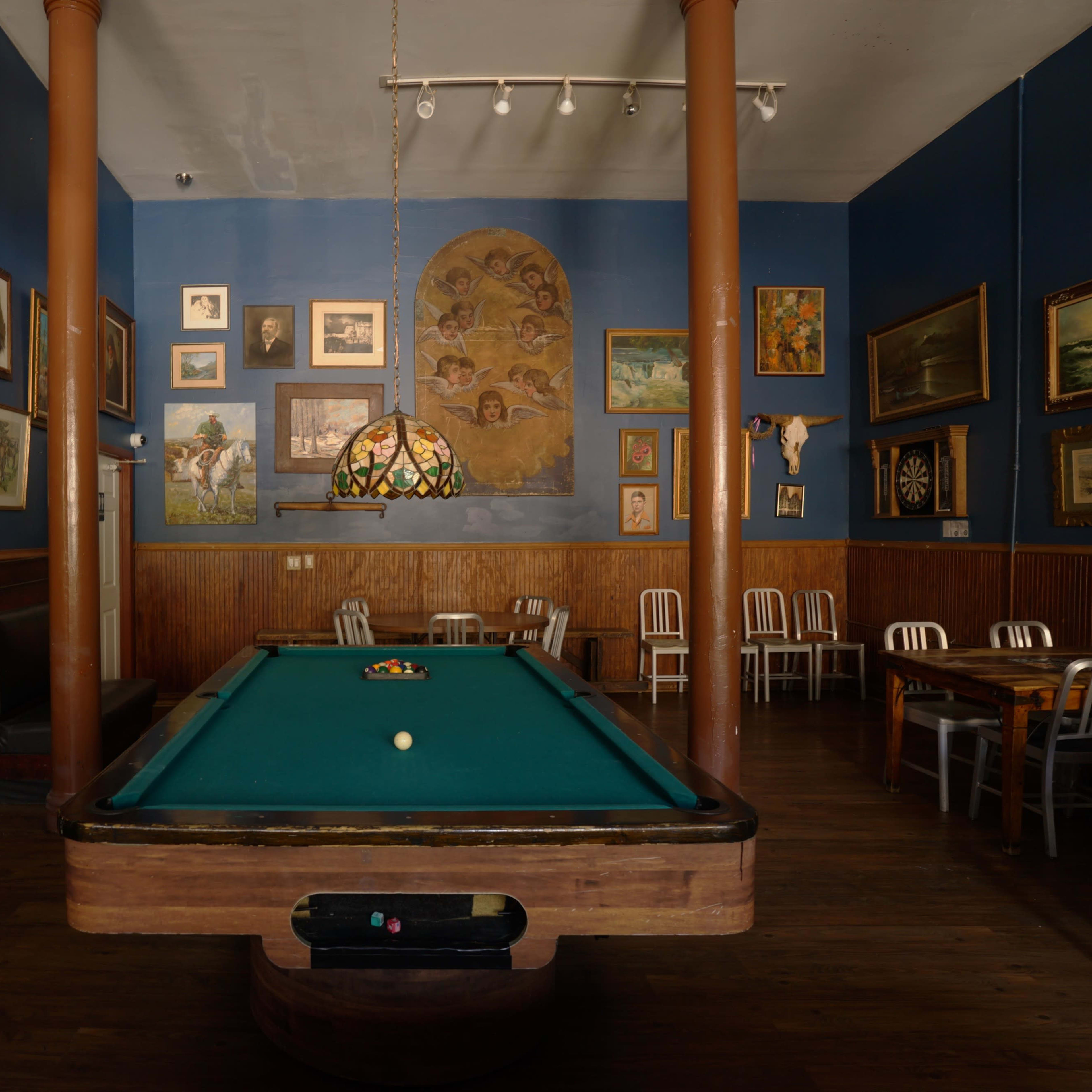 A billiard room featuring a green pool table surrounded by chairs and walls adorned with various framed artworks.