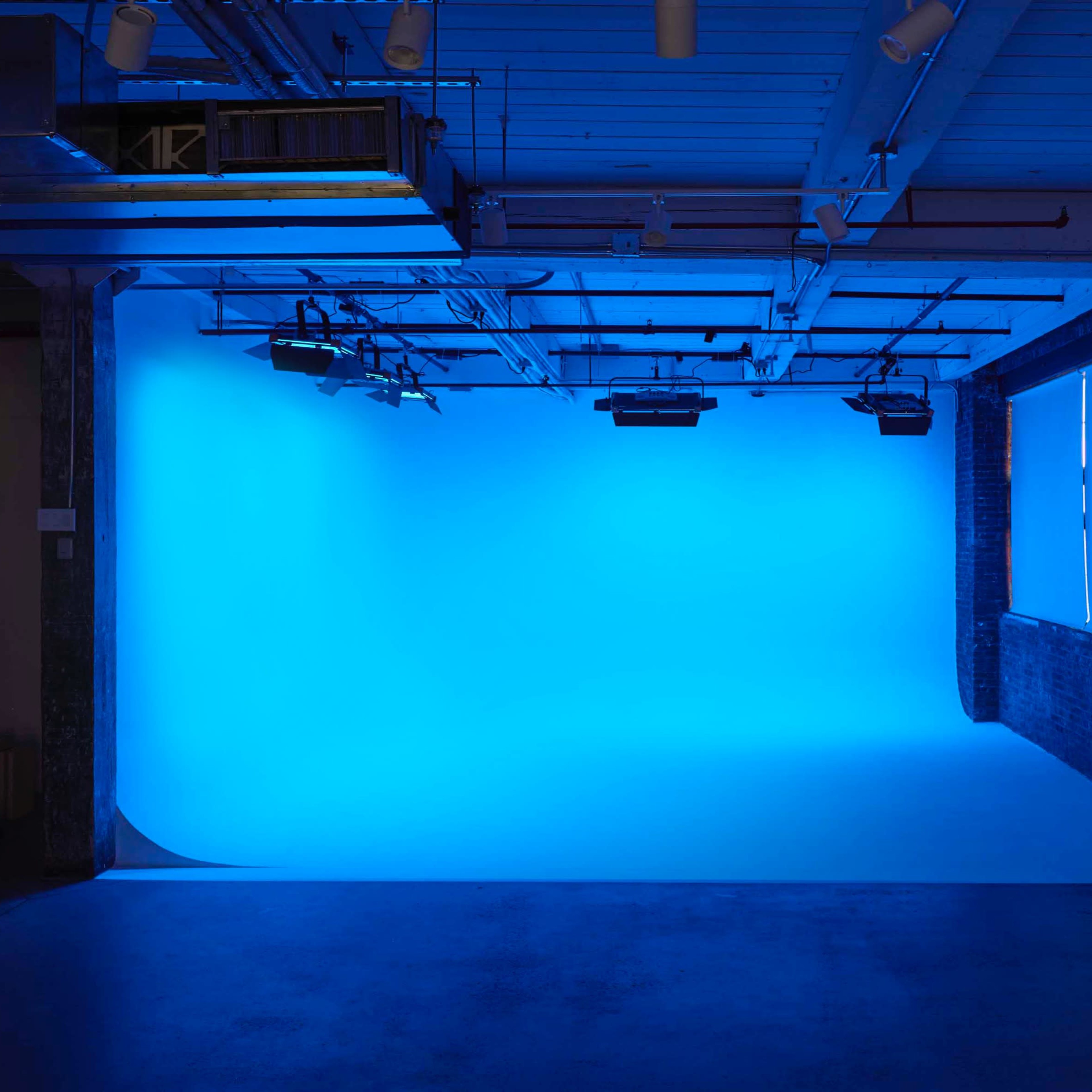 A photography studio featuring a blue backdrop, illuminated by overhead lights and natural light from a window.