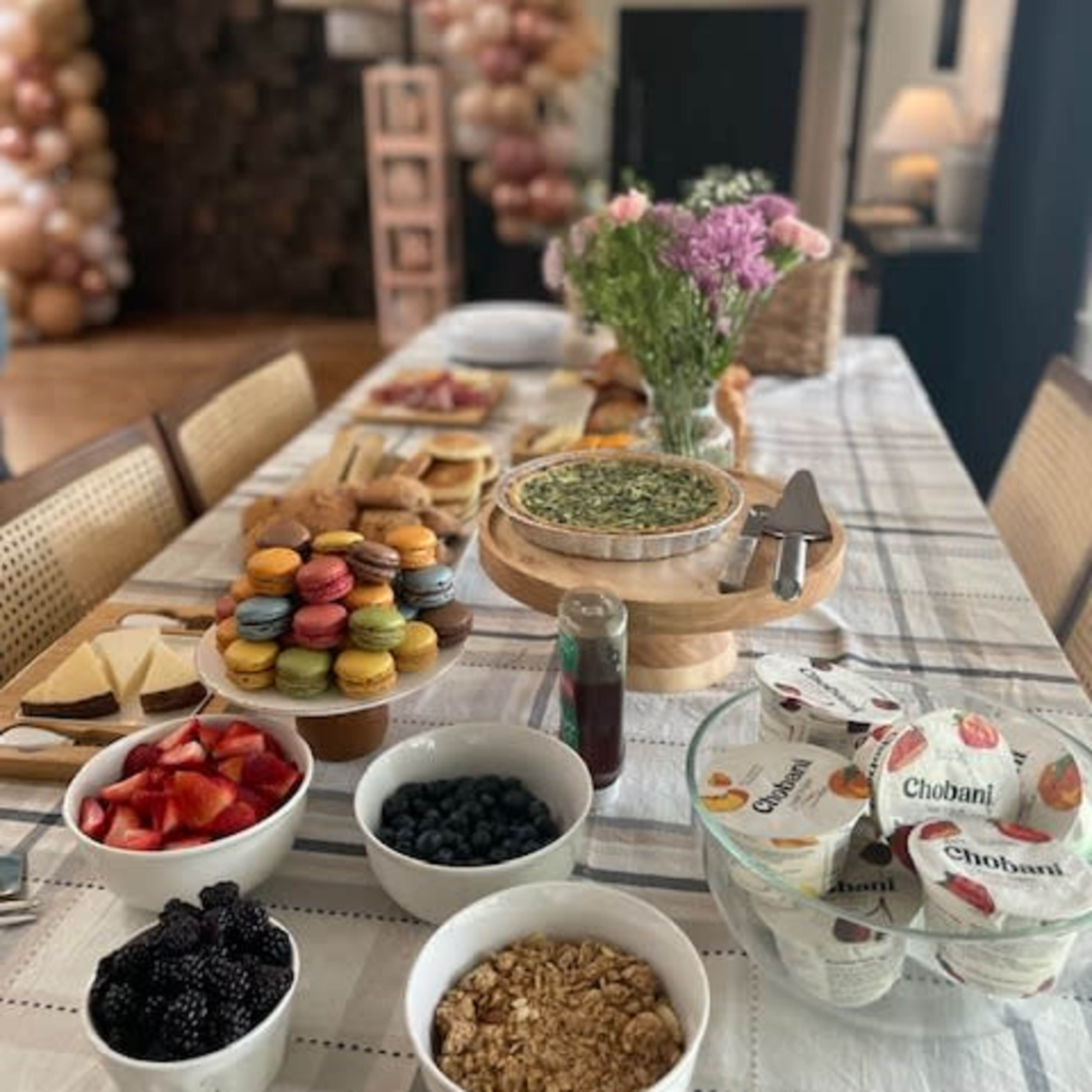 A long table is set with various food items, including fruits, yogurt, pastries, and a cake, in a decorated indoor space.