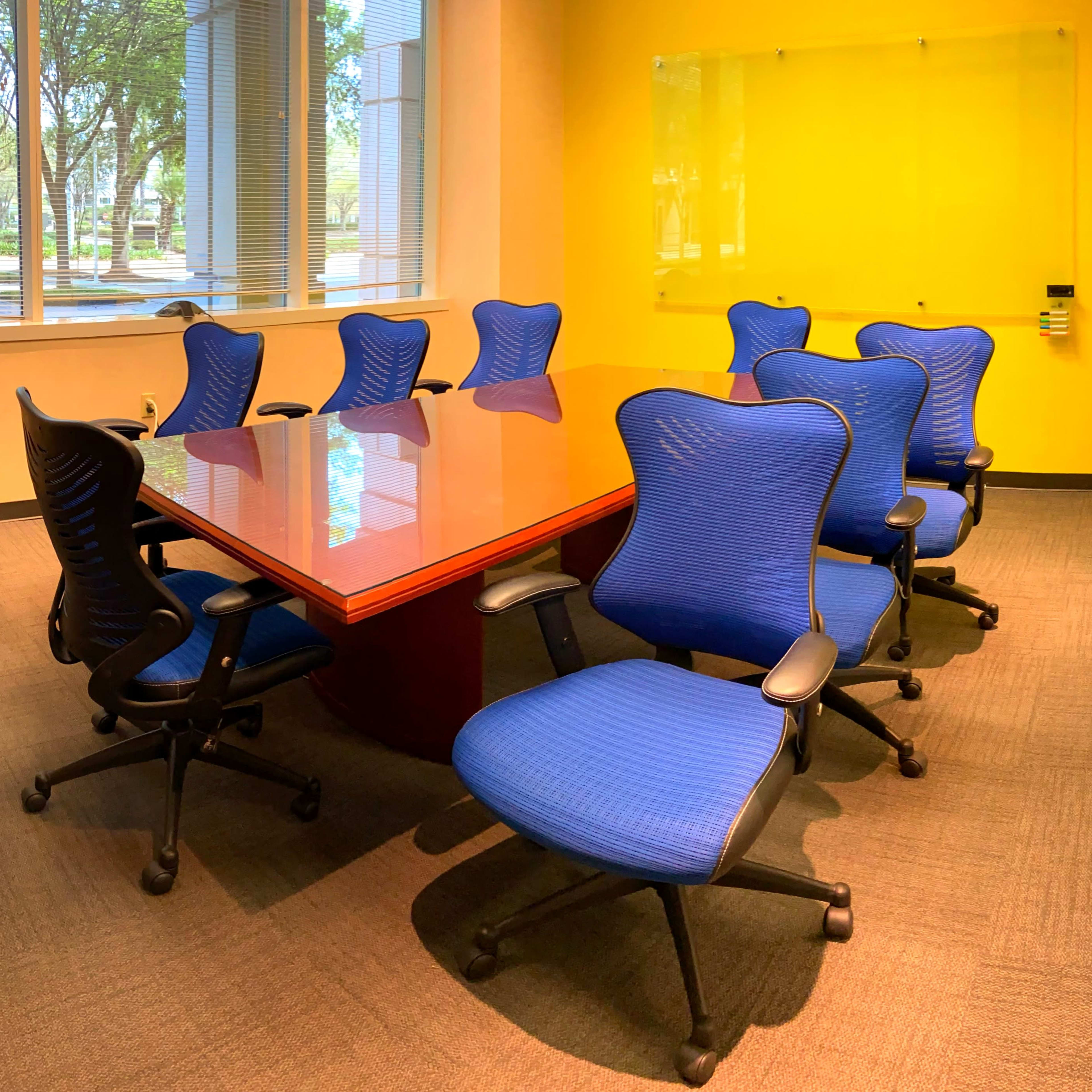 A conference room features a wooden table surrounded by eight blue ergonomic chairs against a bright yellow wall.