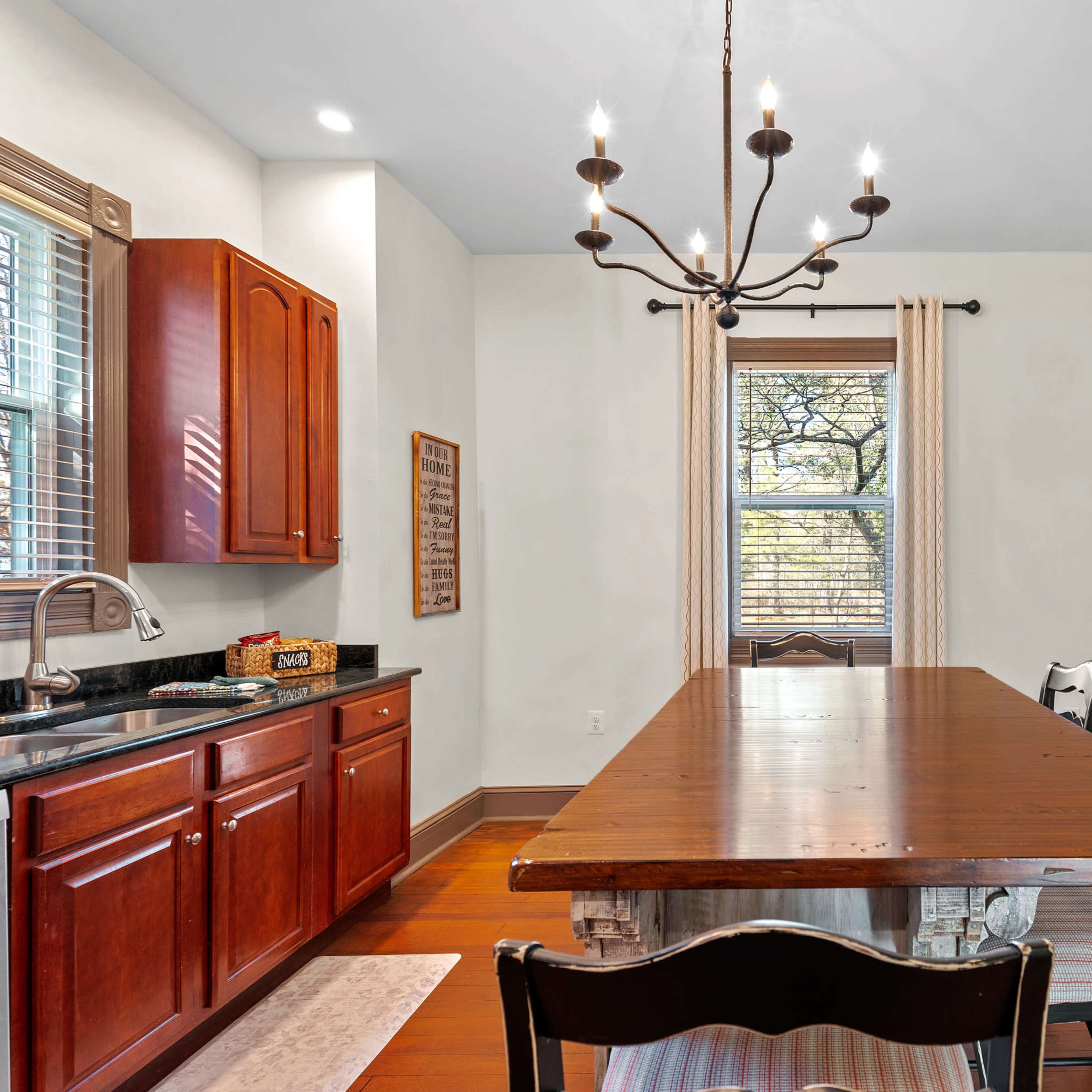 A well-lit dining area features a long wooden table surrounded by chairs, with dark cabinets and a chandelier hanging above.
