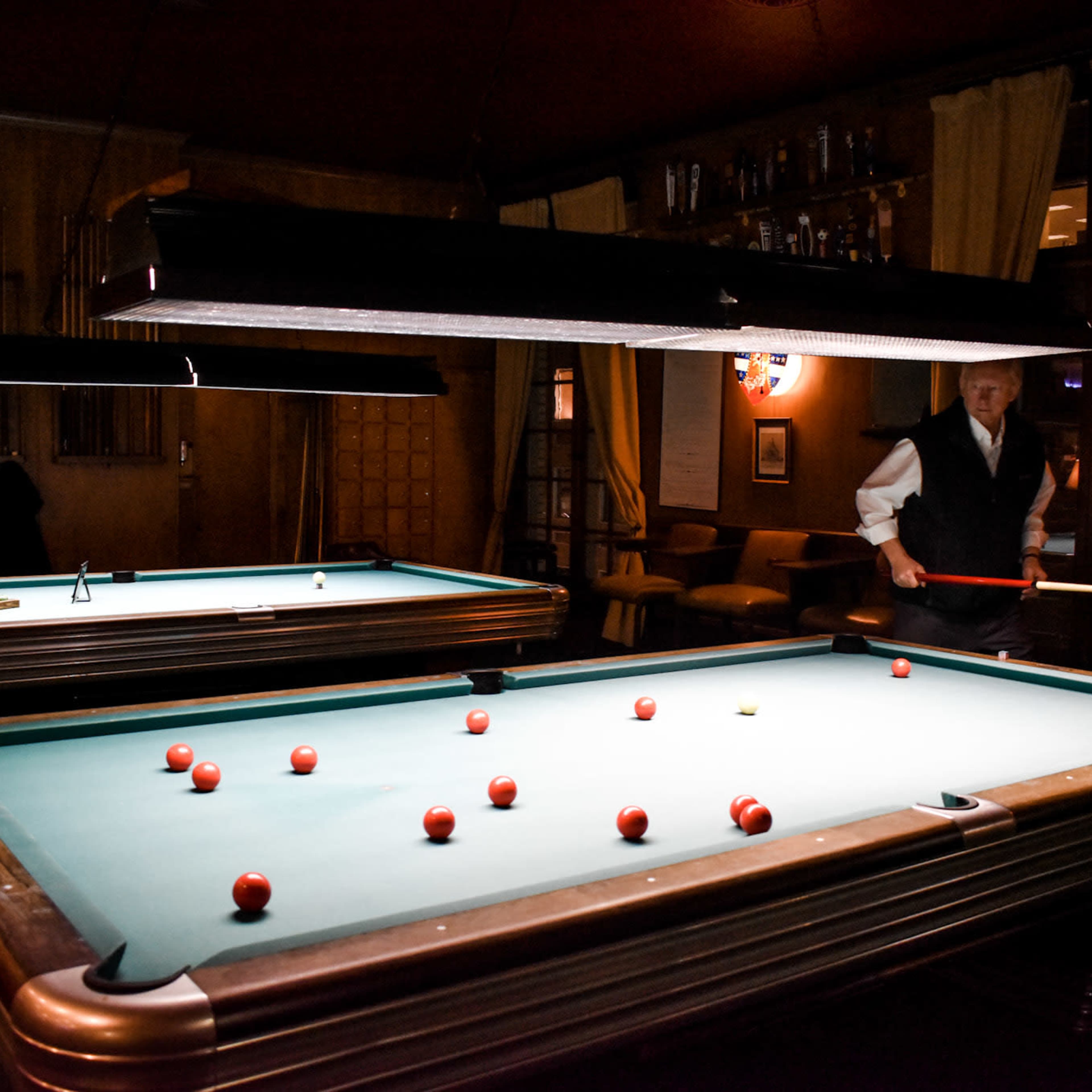 A man stands near a pool table illuminated by overhead lights, with several red balls scattered on the green felt surface.