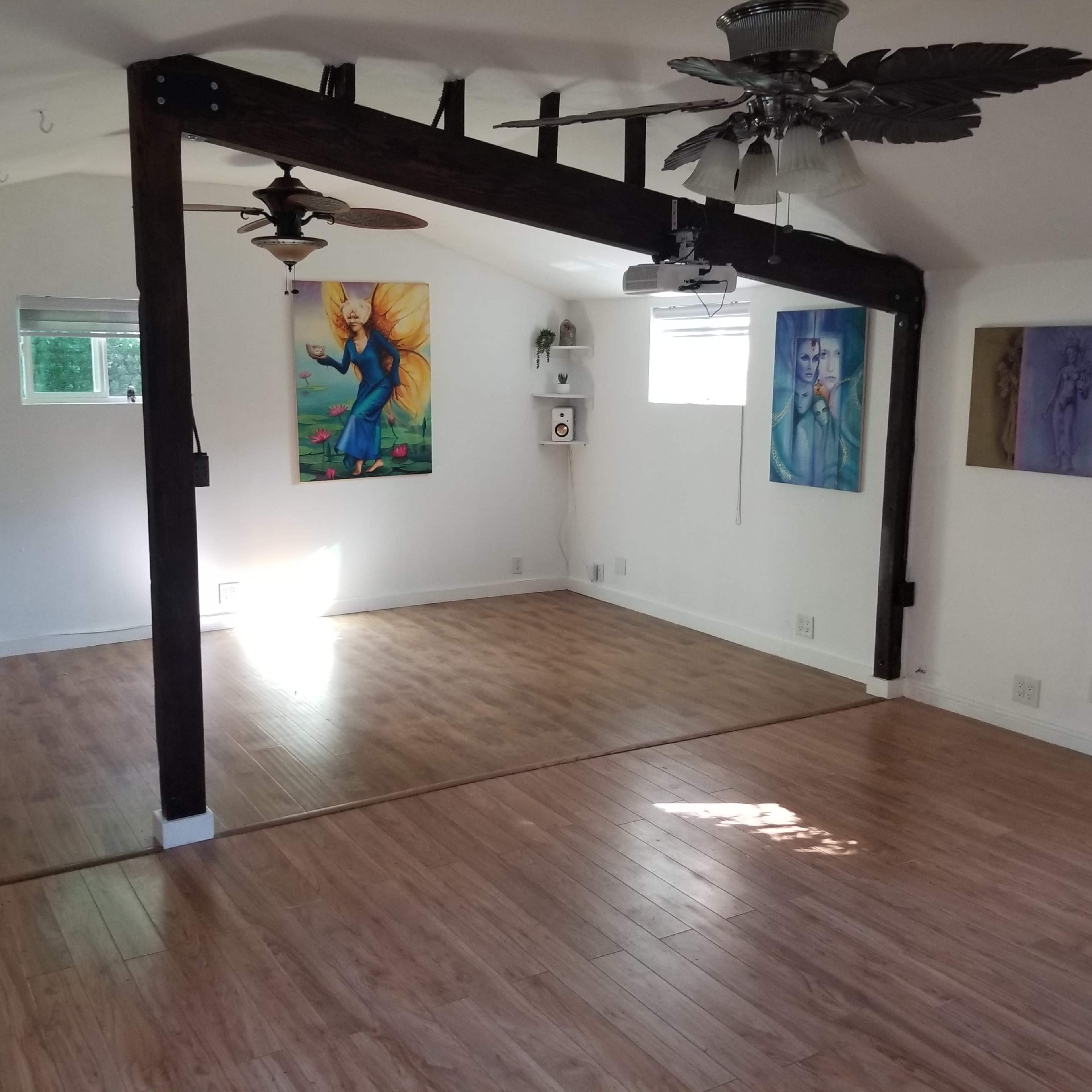 A spacious room with wooden flooring, featuring two decorative wall paintings, two ceiling fans, and a divided area created by a wooden beam.