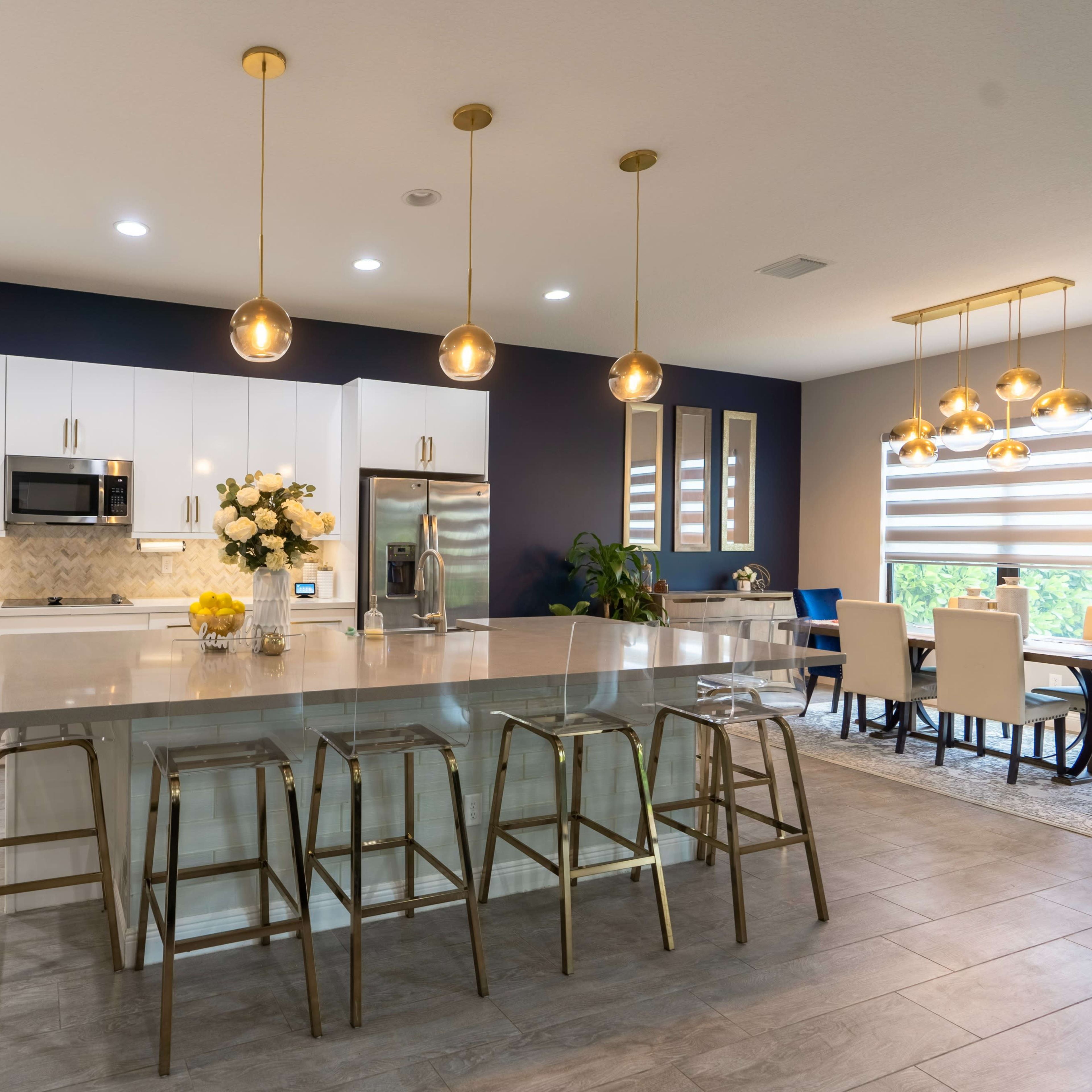 The kitchen features white cabinetry, a large island with golden barstools, and modern pendant lights.