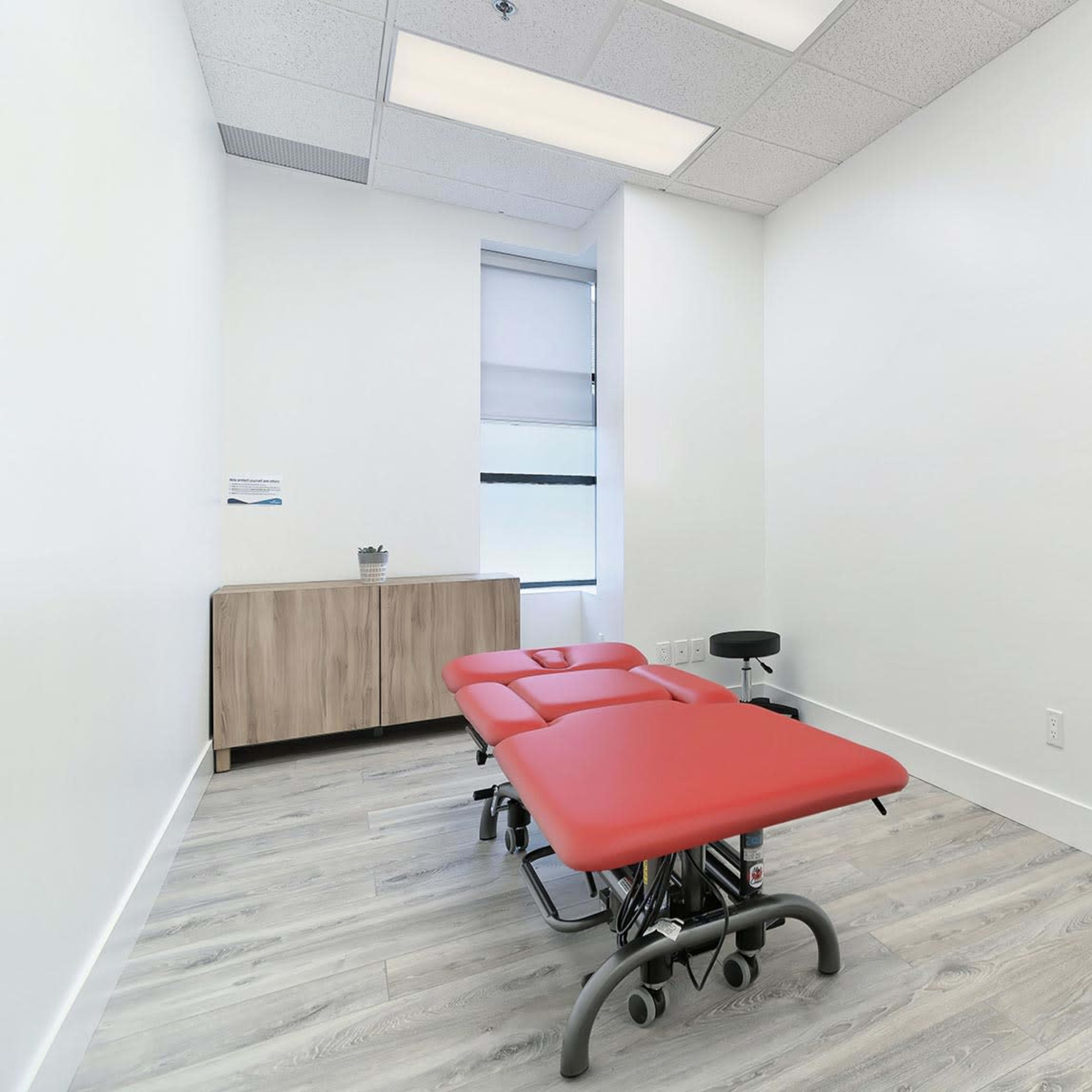 A minimalist treatment room featuring two red examination tables and a wooden cabinet against a white wall.