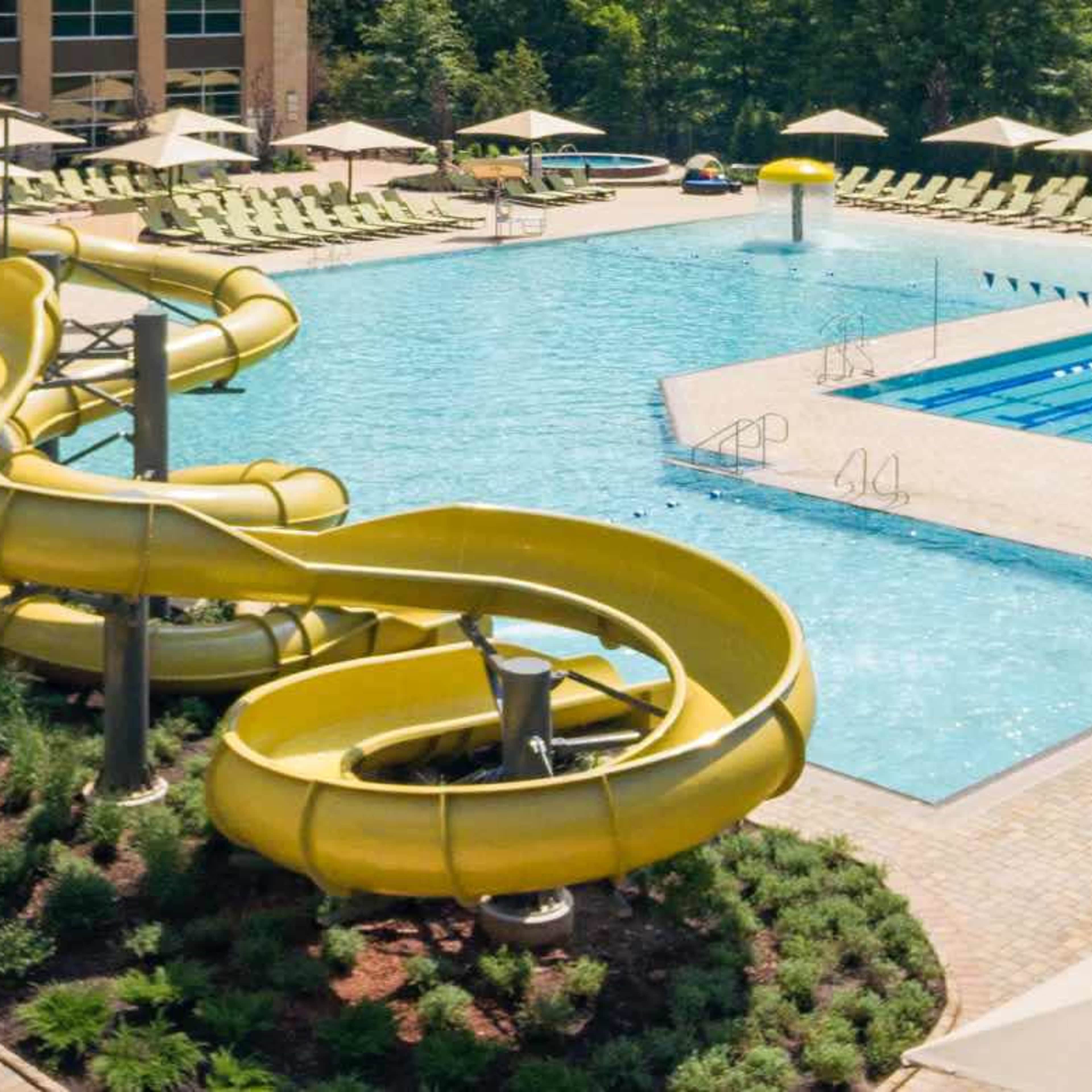 A large swimming pool surrounded by lounge chairs and umbrellas, featuring a yellow water slide on one side.