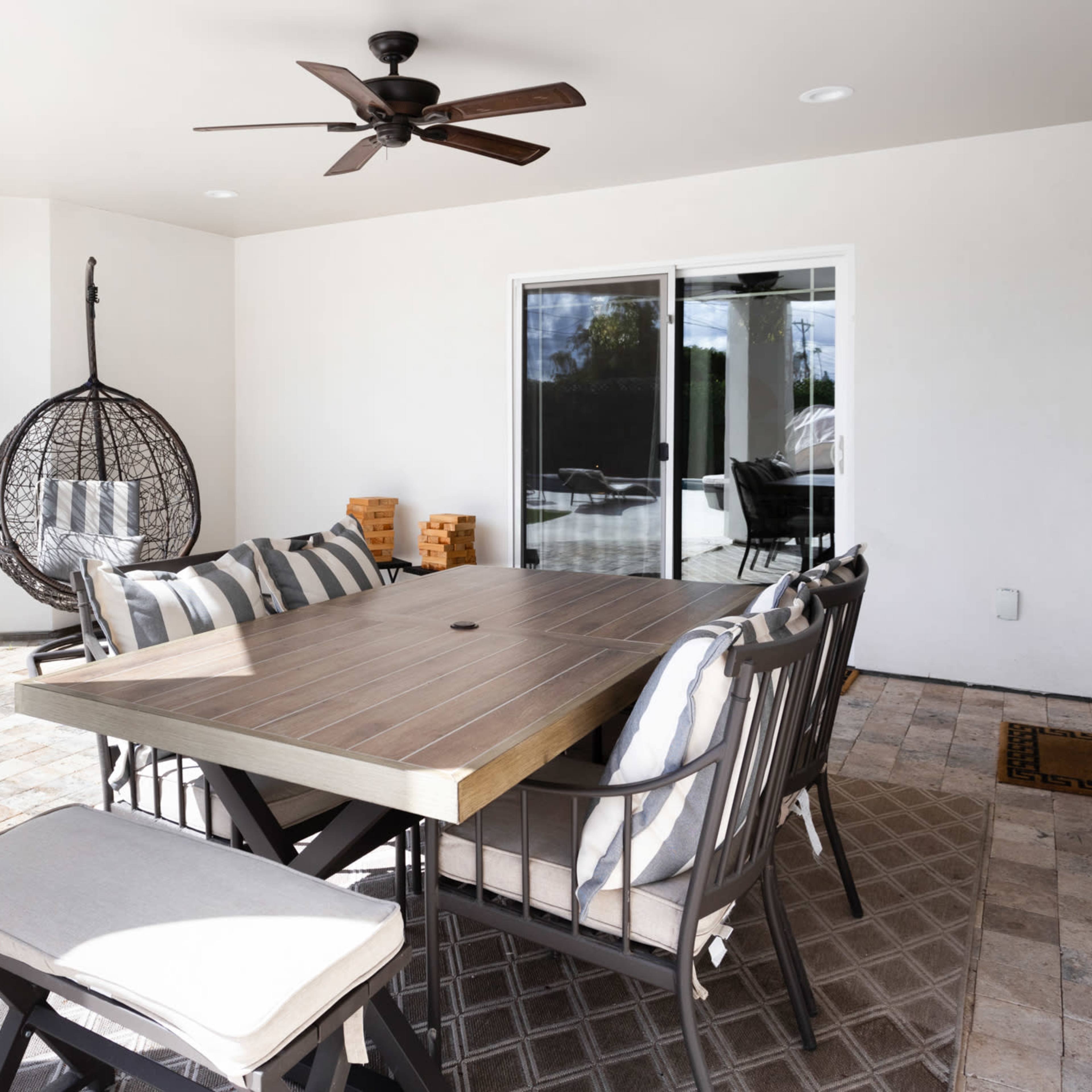 A patio area with a large wooden dining table, several chairs, a hanging chair, and a ceiling fan, all set against a backdrop of white walls.