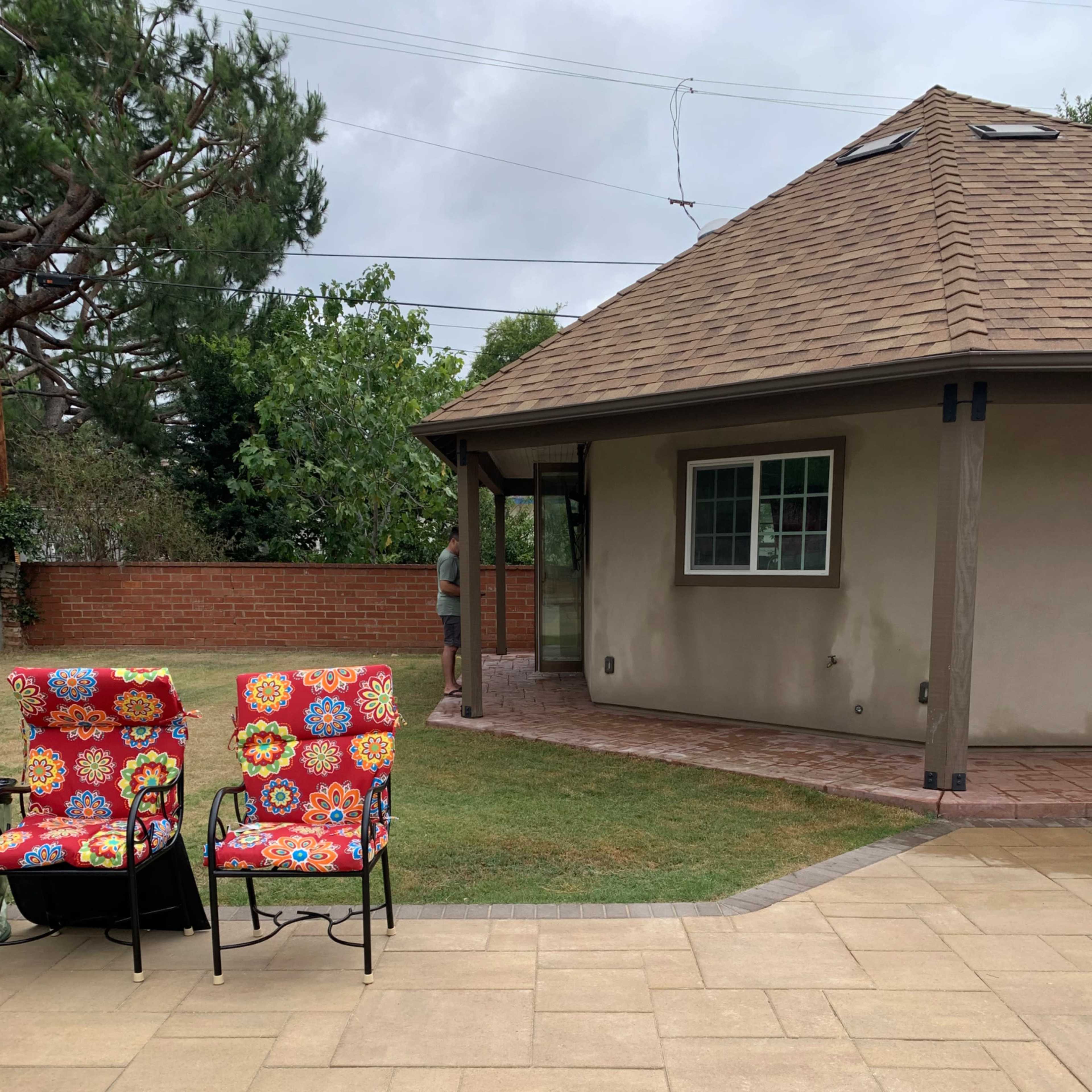 A backyard area with two patterned chairs facing a garden space, a brick walkway, and a house in the background.