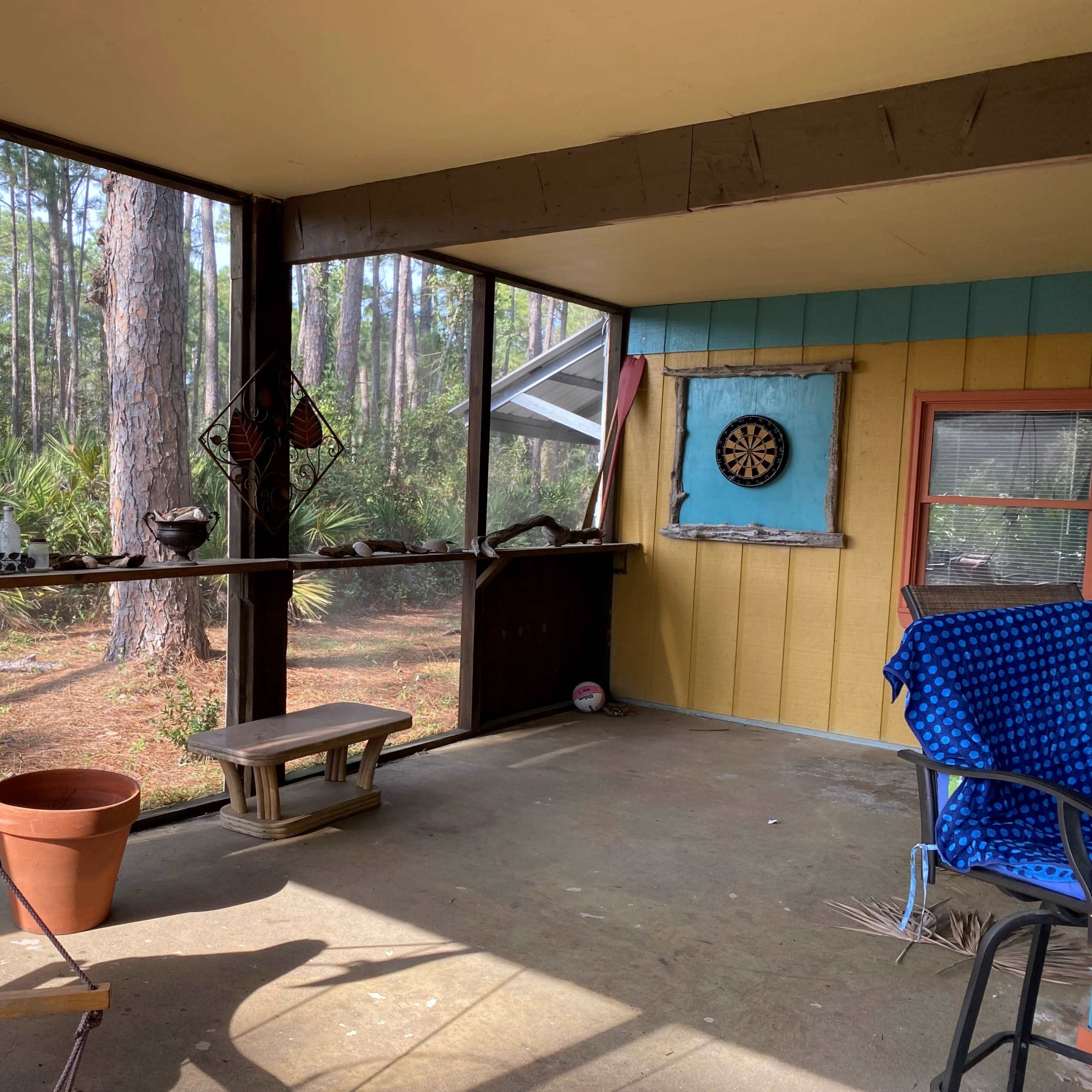 A screened-in porch with a dartboard mounted on the wall, a wooden bench, and a colorful chair, surrounded by trees in the background.