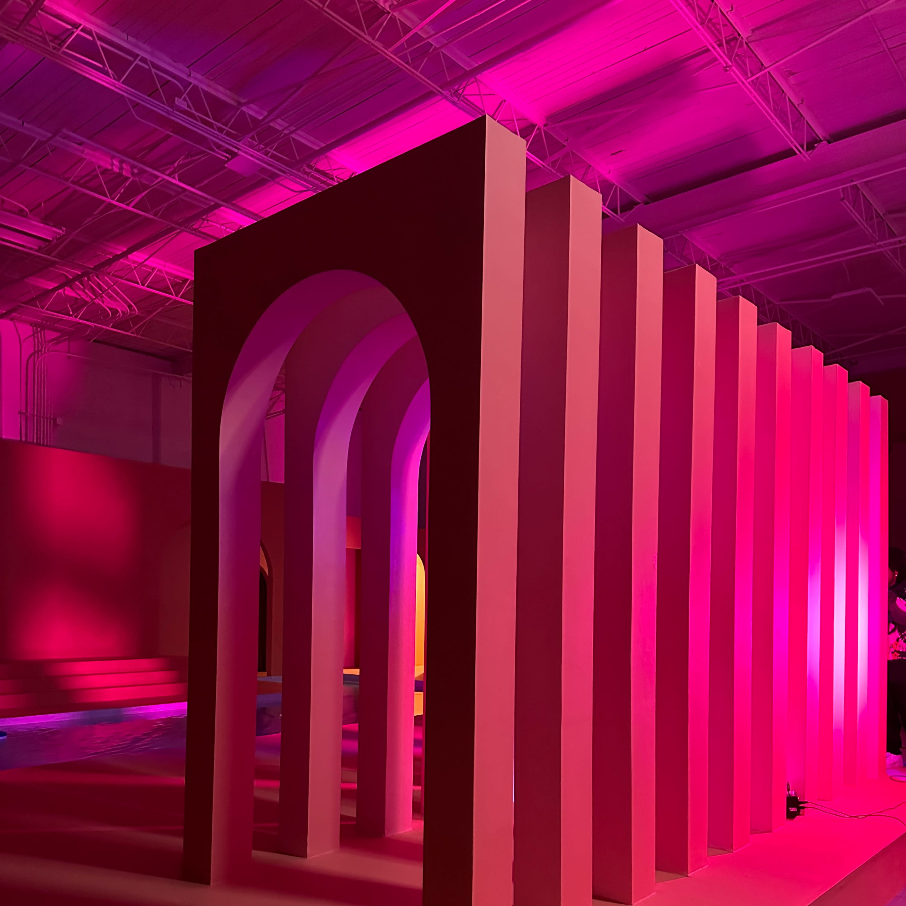 A series of large, colorful arches and vertical structures illuminated by pink lighting in an industrial space.