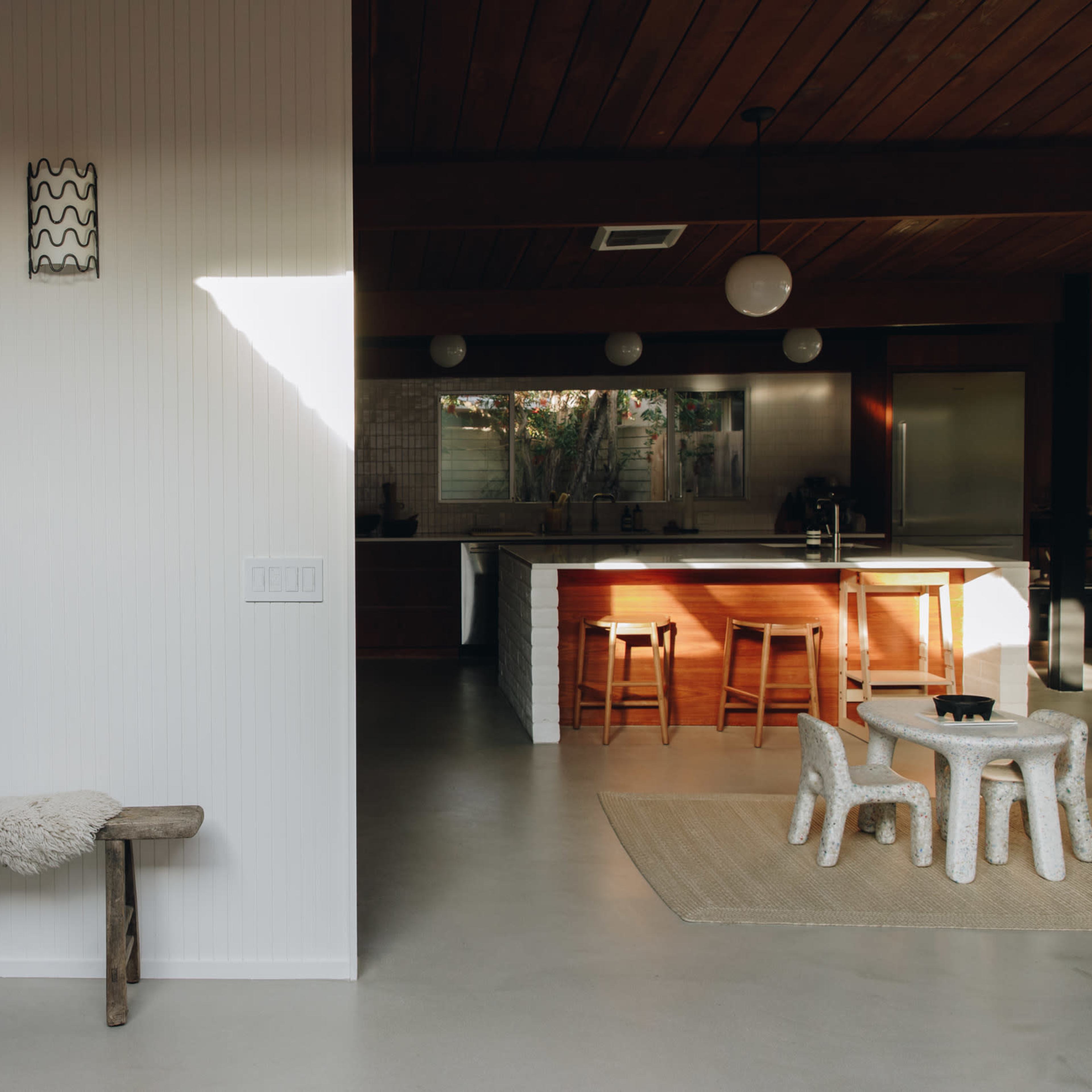A minimalist interior features a wooden bench and a white wall on one side, with a kitchen and dining area visible in the background.