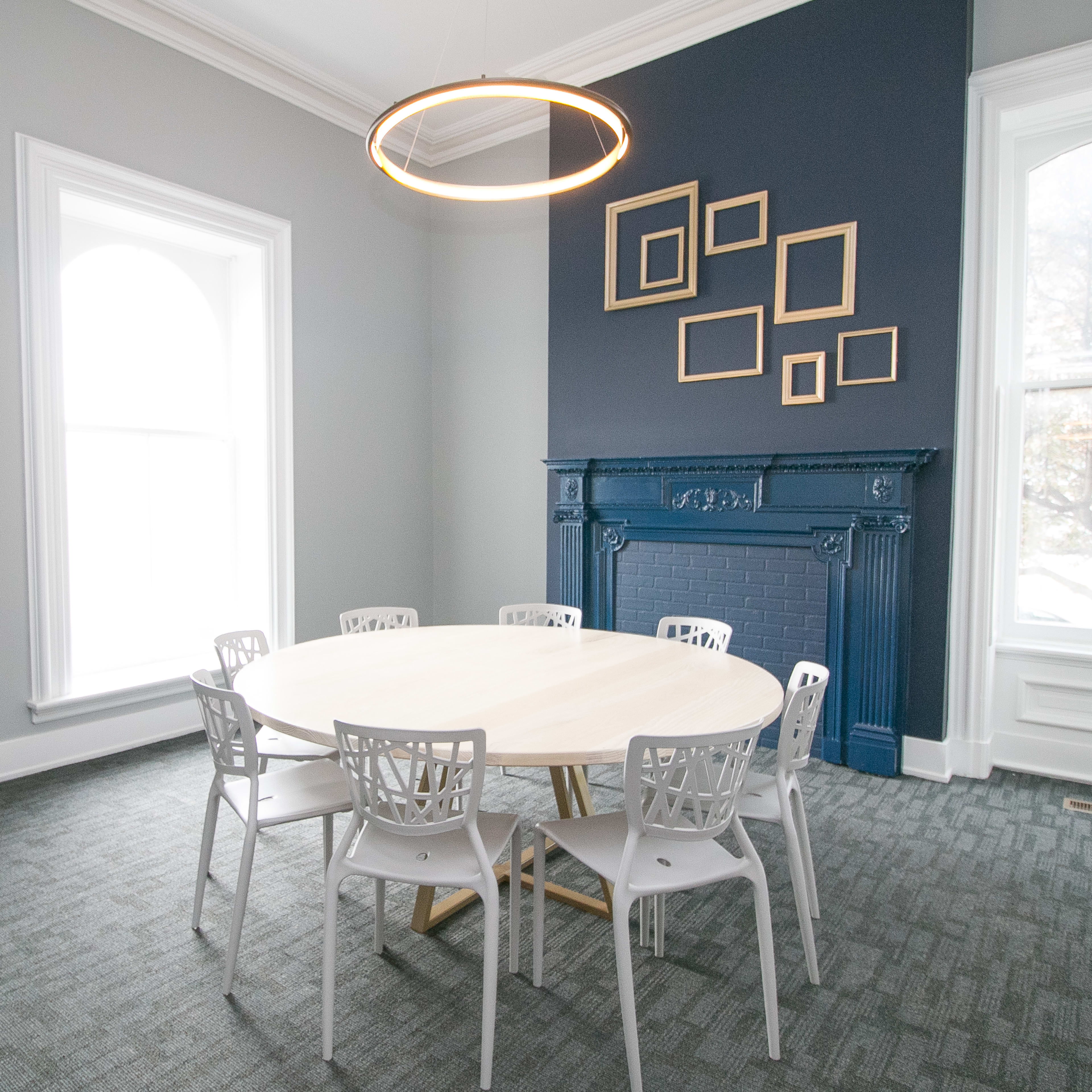A round wooden table is surrounded by six white chairs in a light-filled room featuring a dark blue wall with framed pictures and a decorative fireplace.
