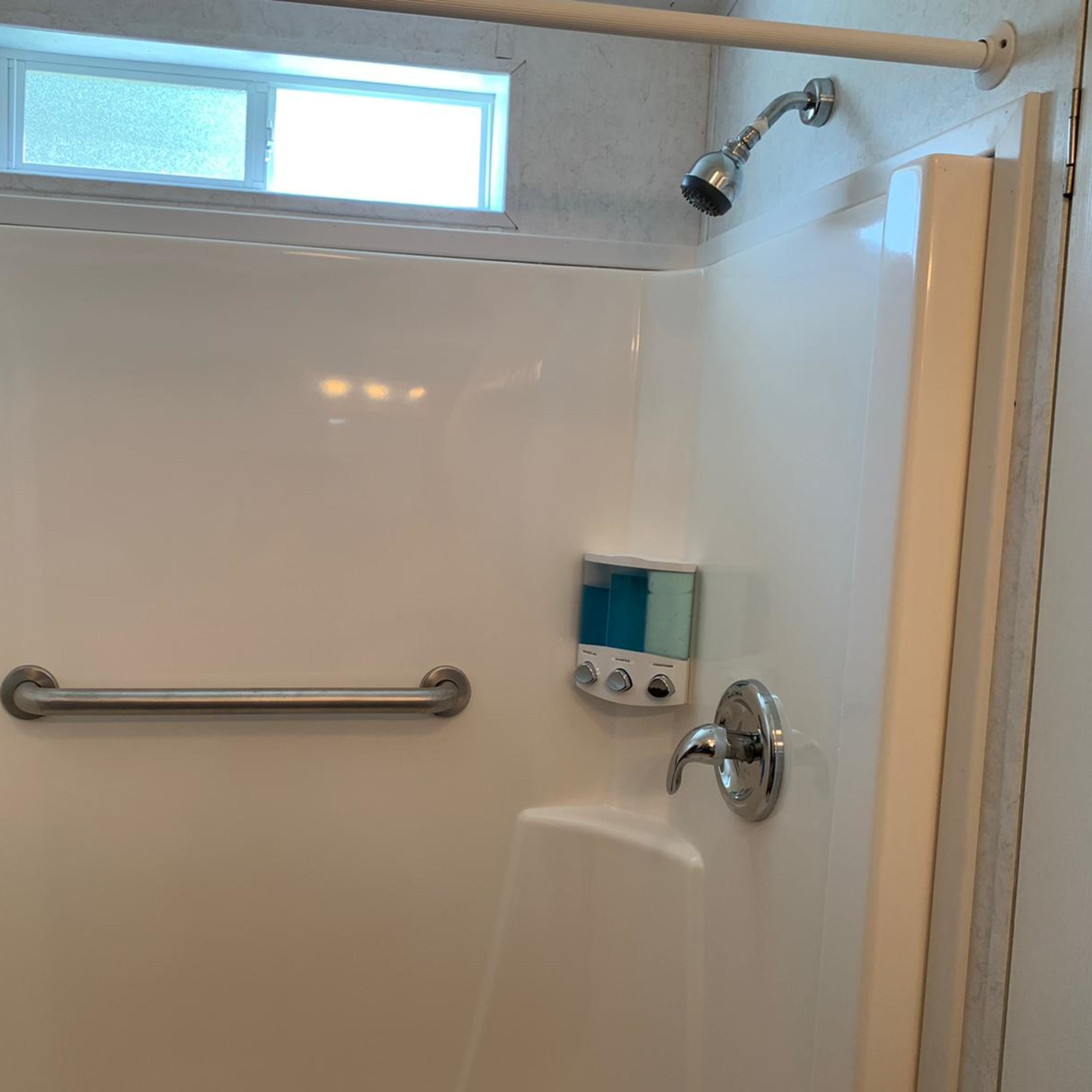 A clean, white shower stall with a showerhead, grab bar, and a soap dispenser mounted on the wall.
