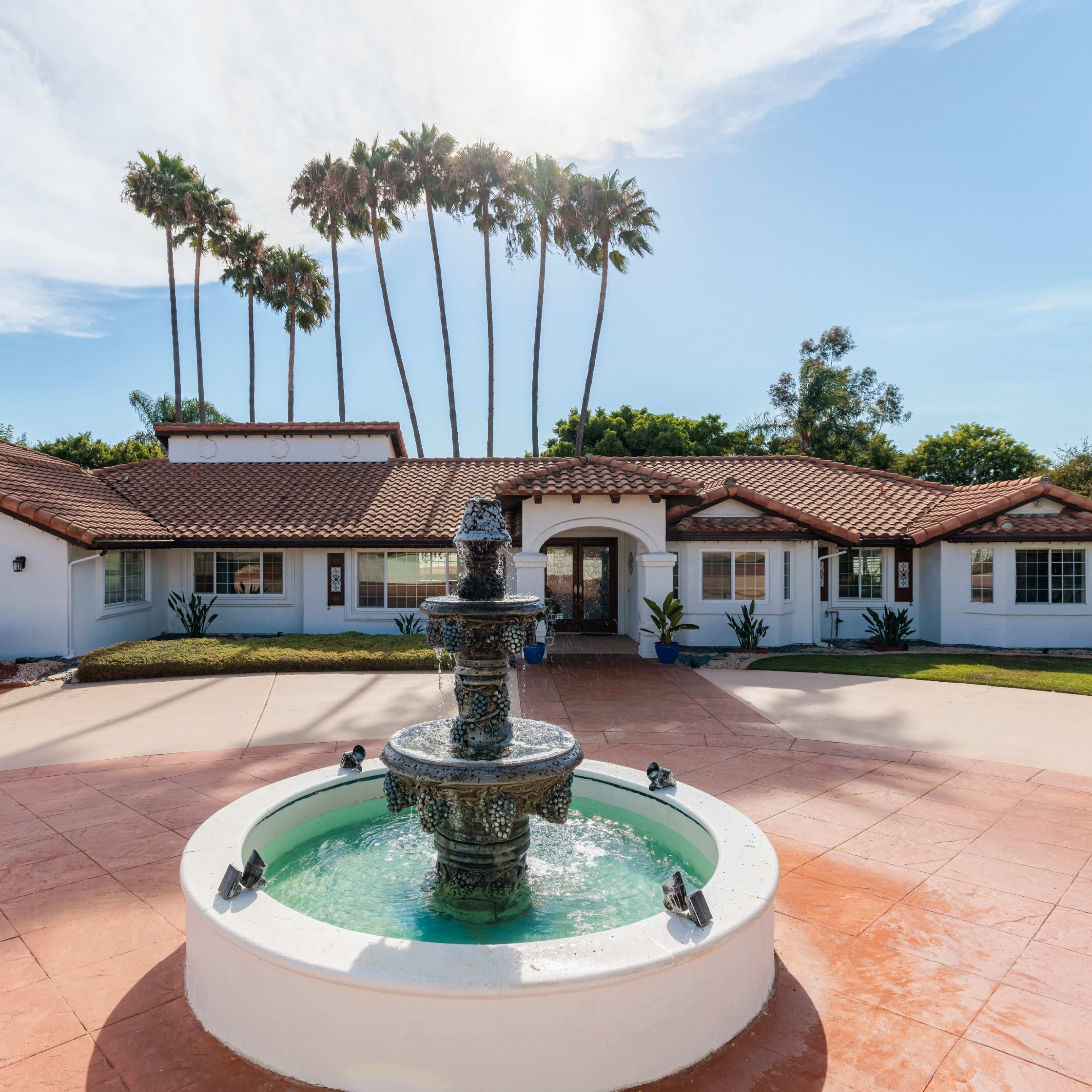 A fountain is centered in a landscaped courtyard in front of a large, single-story house with a red-tiled roof and palm trees in the background.