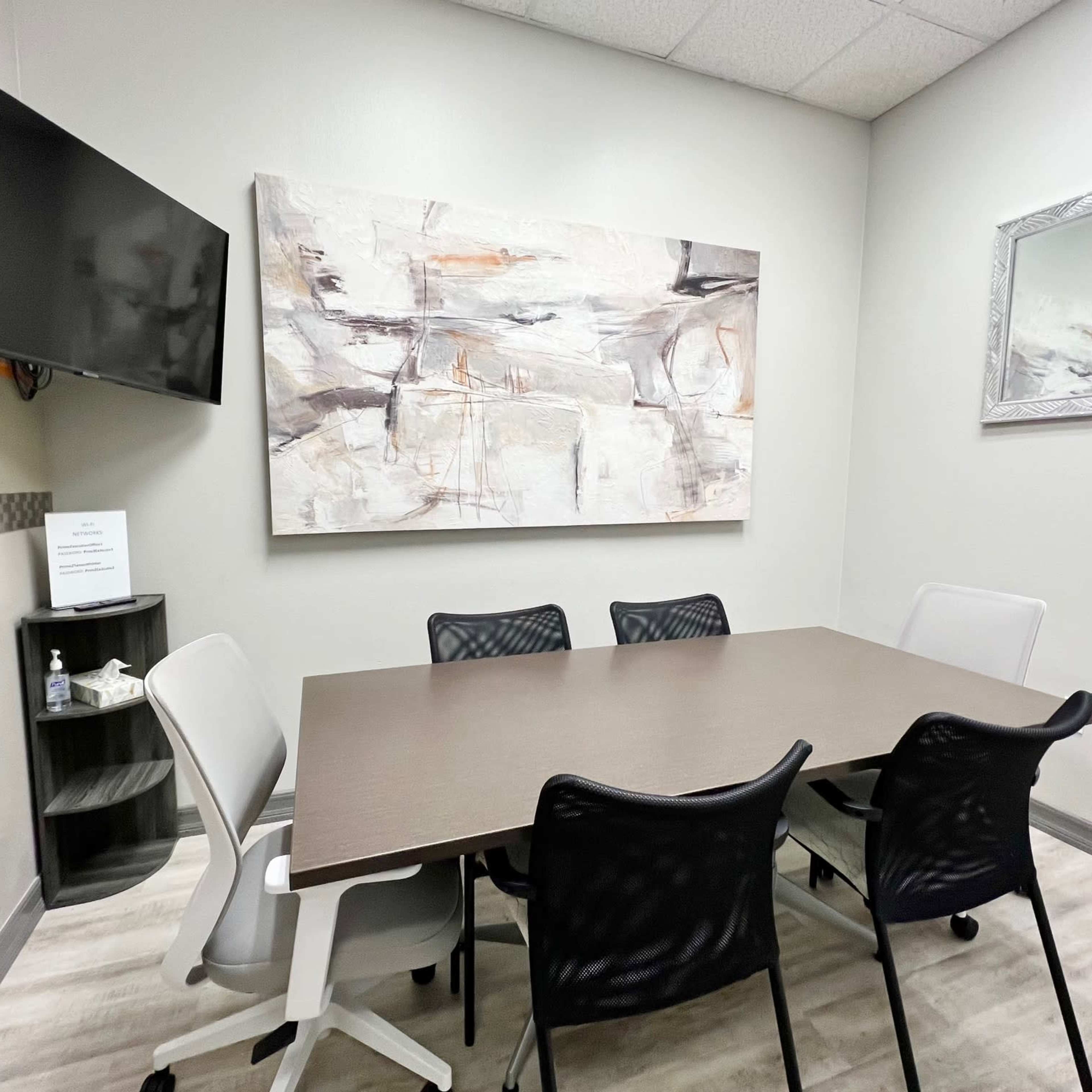 A small meeting room with a rectangular table surrounded by six chairs, a wall-mounted TV, and abstract artwork on the wall.