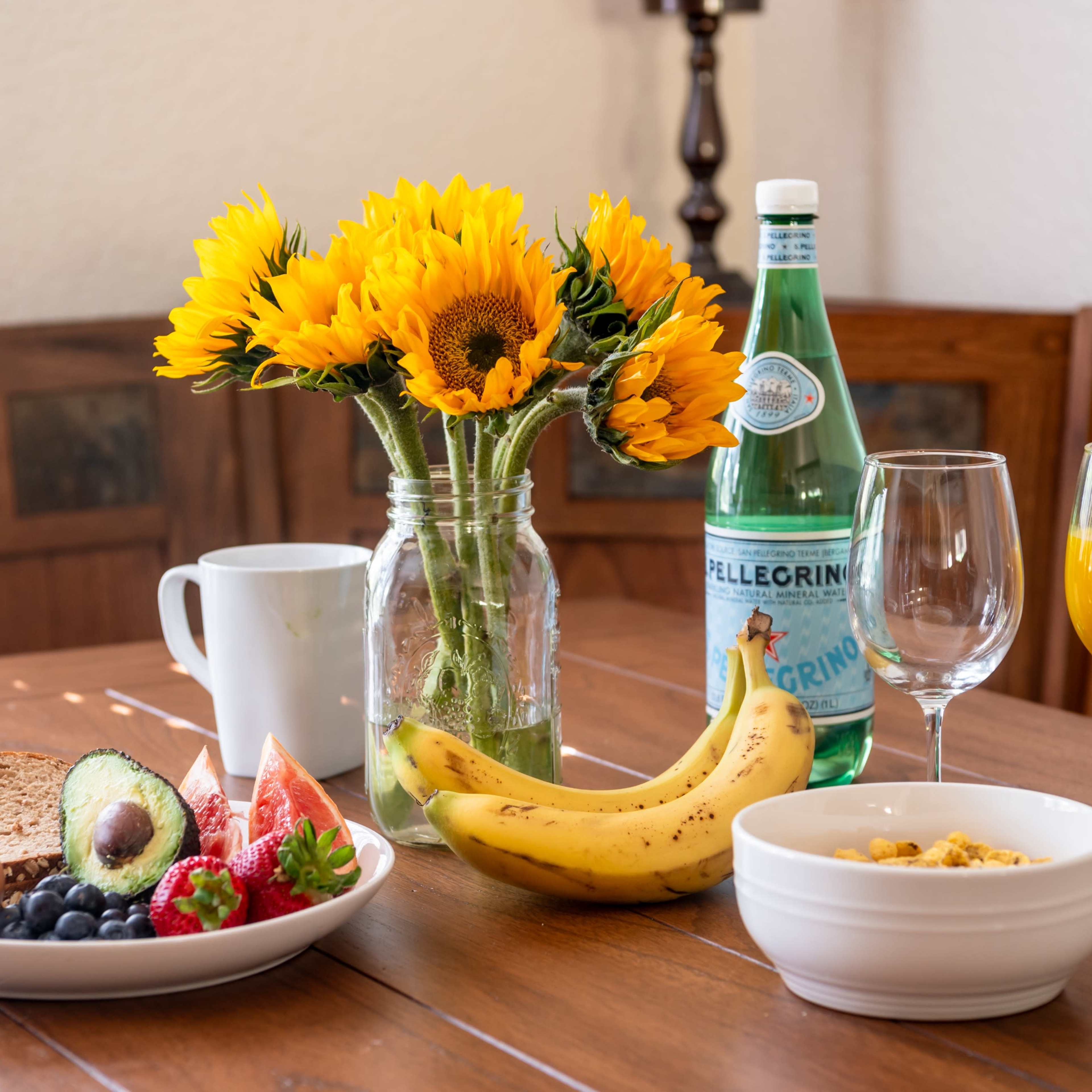 A breakfast table is set with two eggs, a bowl of cereal, assorted fruits, a glass of orange juice, sparkling water, and a vase of sunflowers.