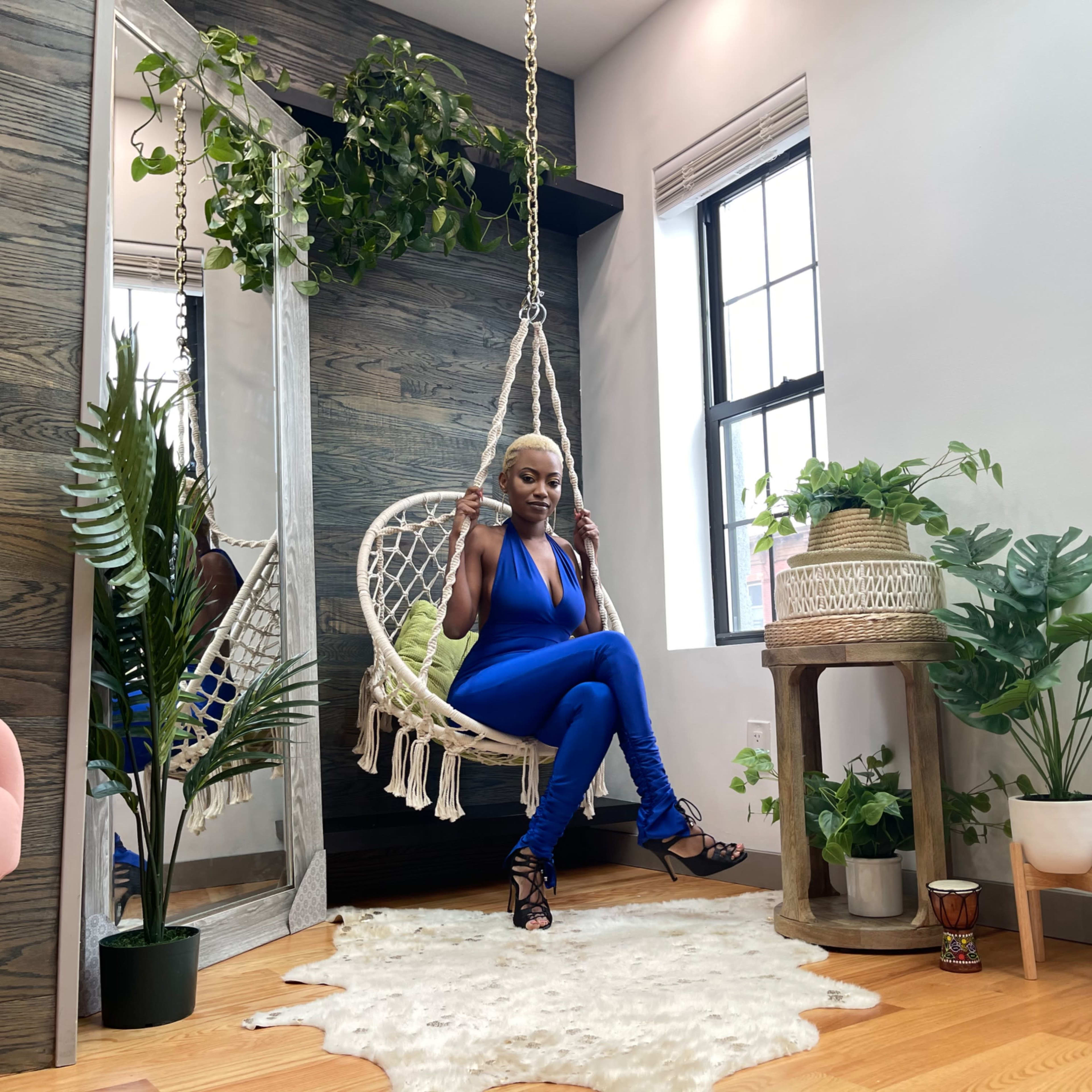 A person sits on a hanging macramé chair surrounded by various indoor plants in a well-lit room with wooden accents.