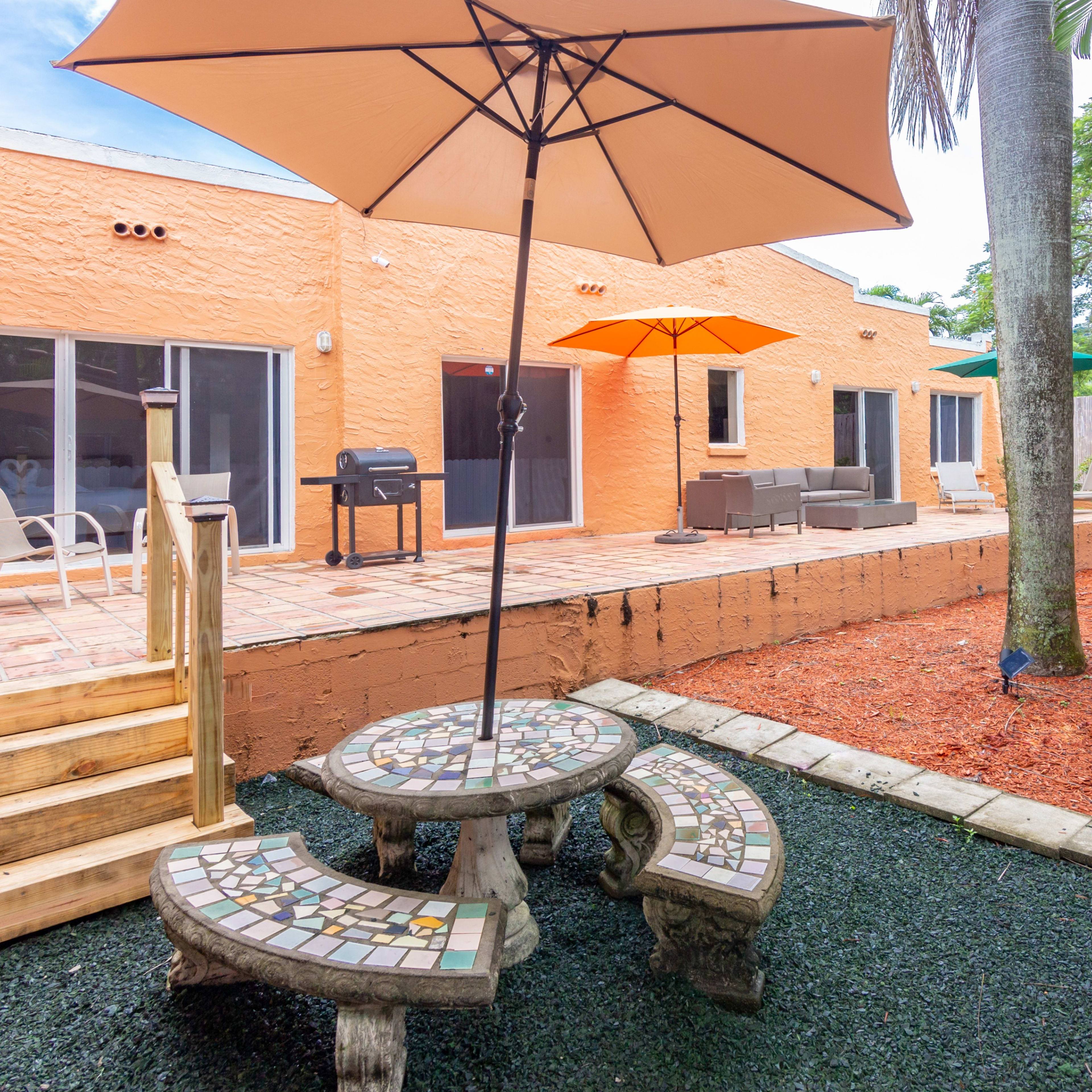 A backyard patio area with a mosaic table and seating, surrounded by mulch and grass, with a grill and lounge furniture against a colorful wall.
