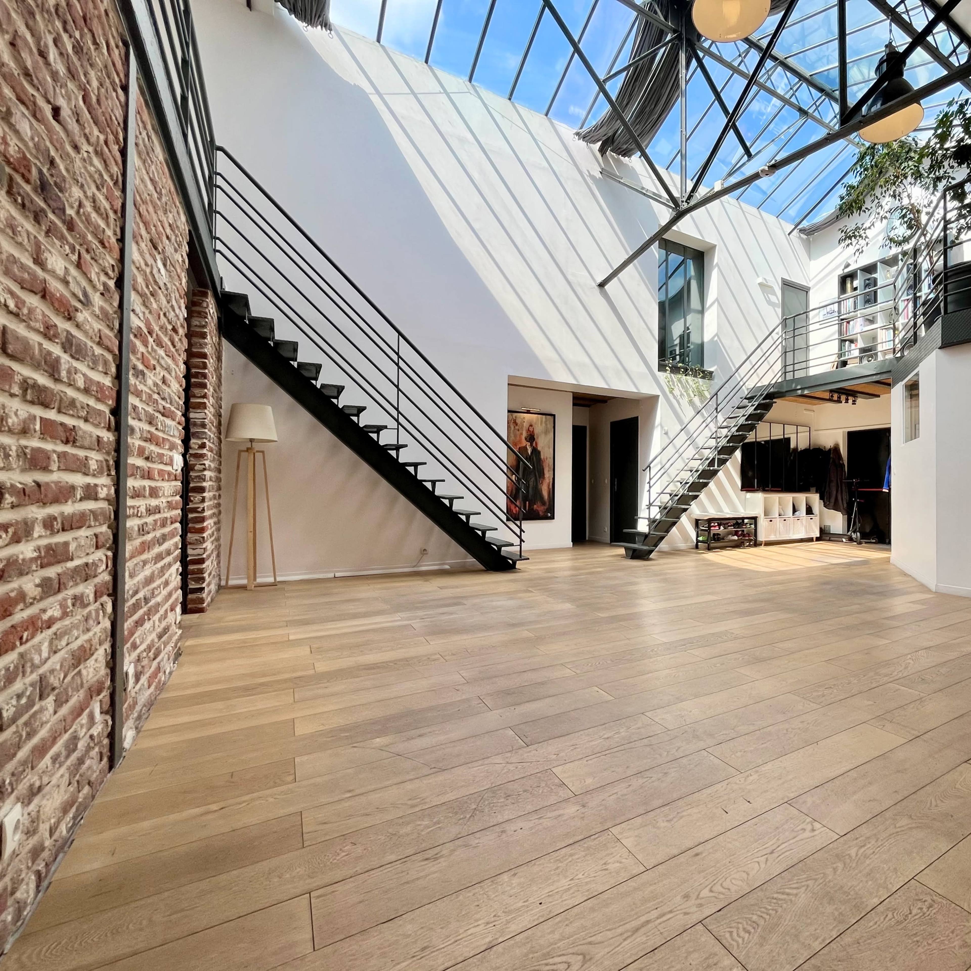 A spacious interior with wooden flooring, a double staircase, large windows allowing natural light, and a mix of modern and industrial design elements.