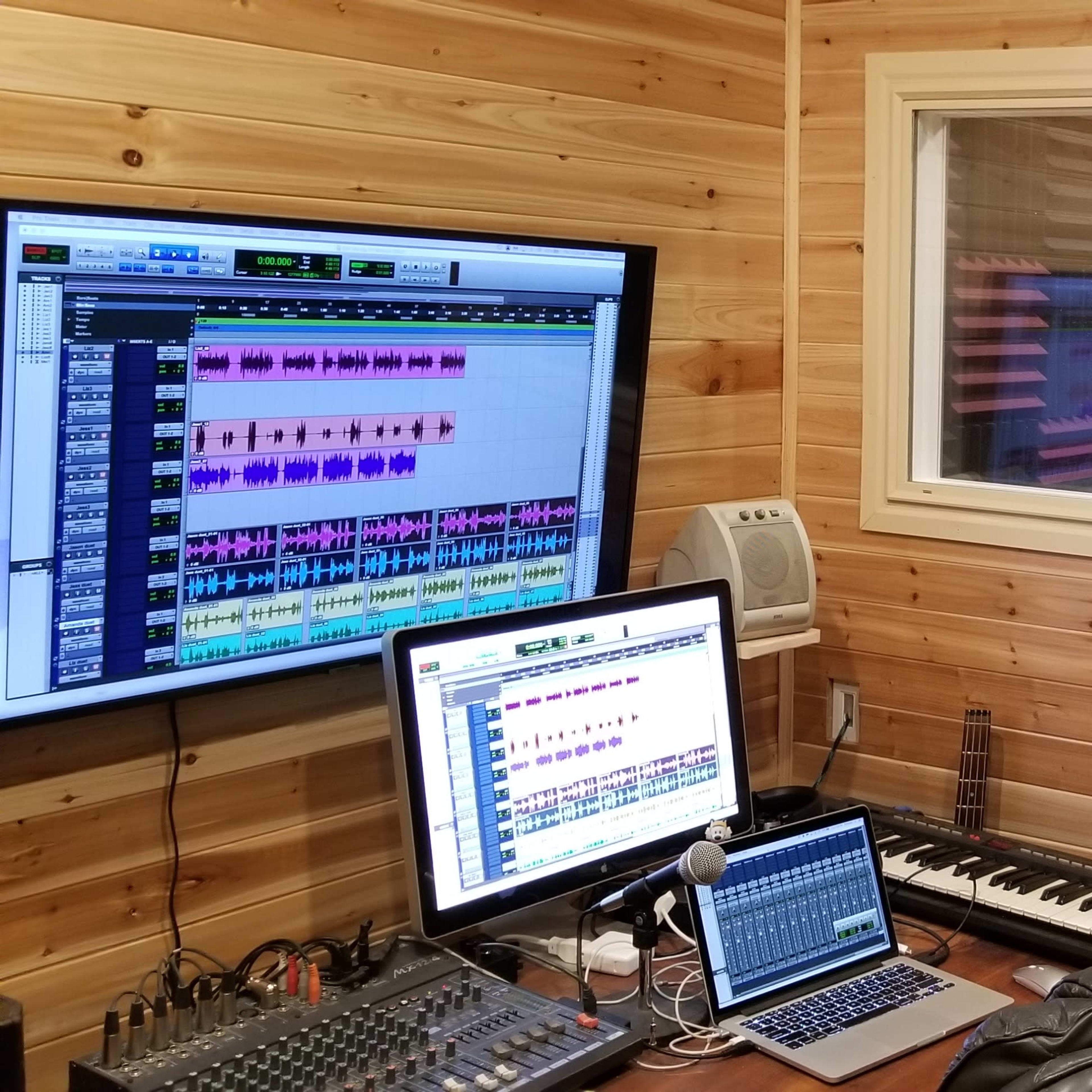 A sound recording studio with a computer setup, a keyboard, and a vocal booth in the background.