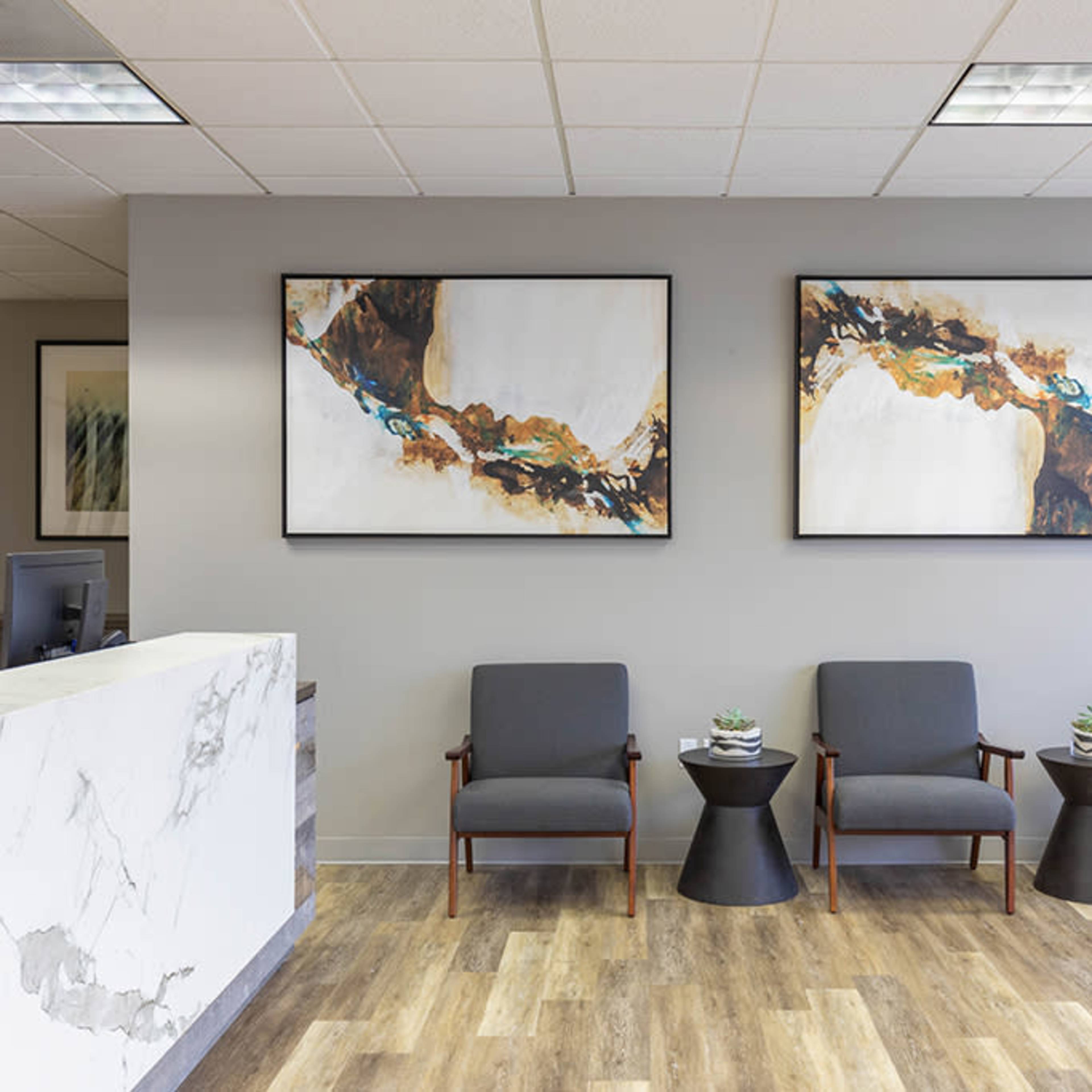 A modern waiting area featuring three gray chairs, small side tables, and two abstract paintings on the wall.