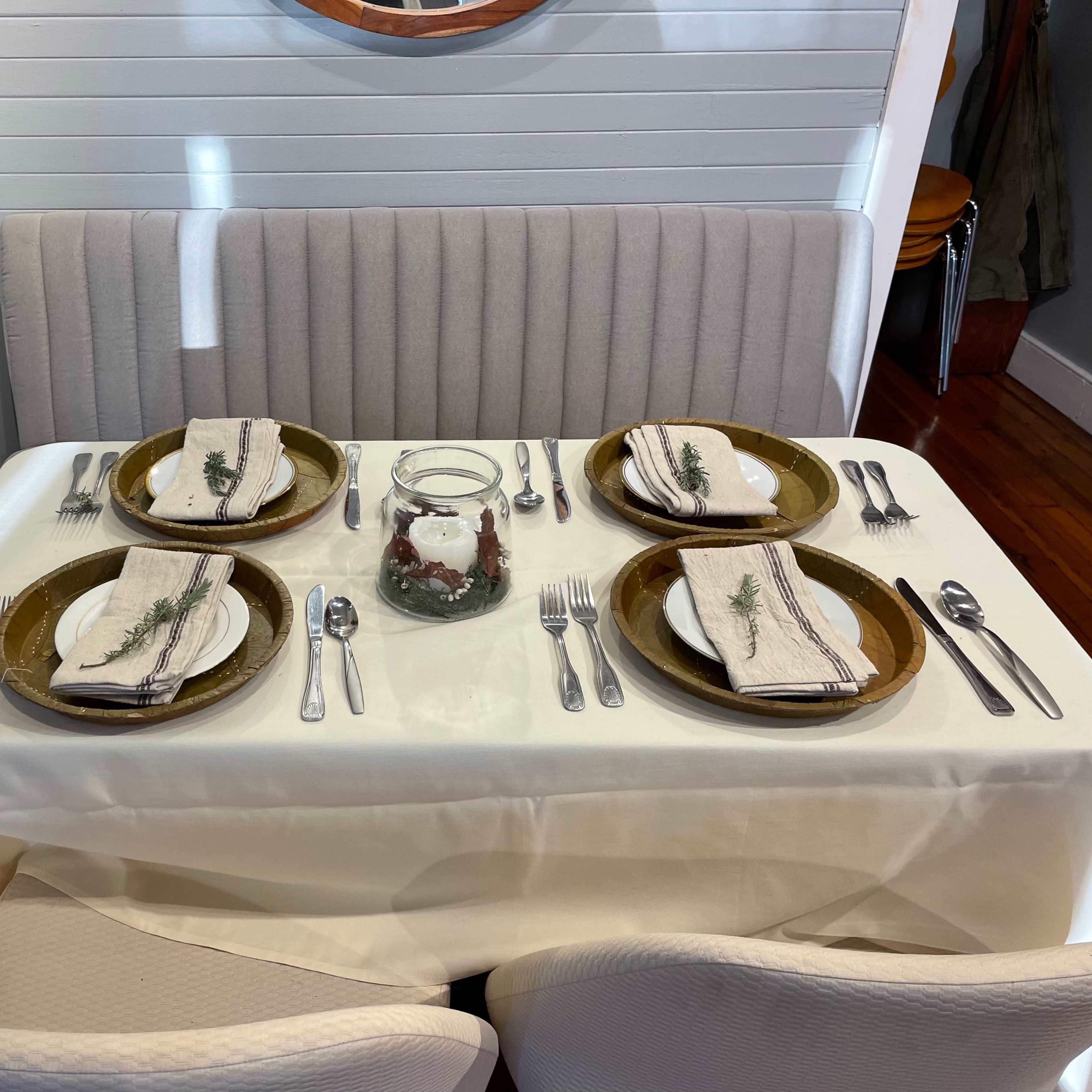 A neatly set dining table with four place settings, featuring plates, cutlery, and decorative napkins, is positioned beside a booth against a wall.