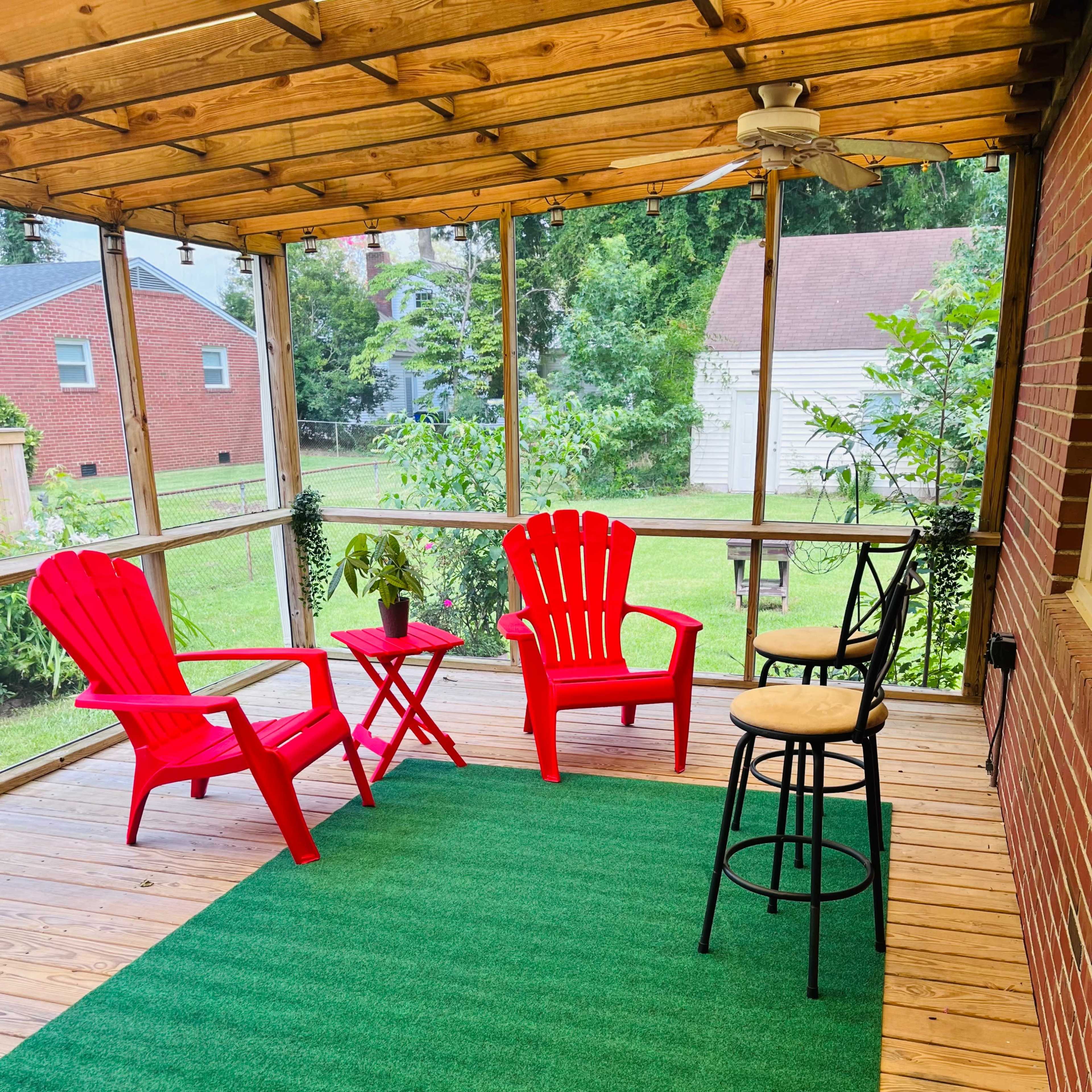 A screened porch featuring two red adirondack chairs, a small table, a black stool, and a green rug.