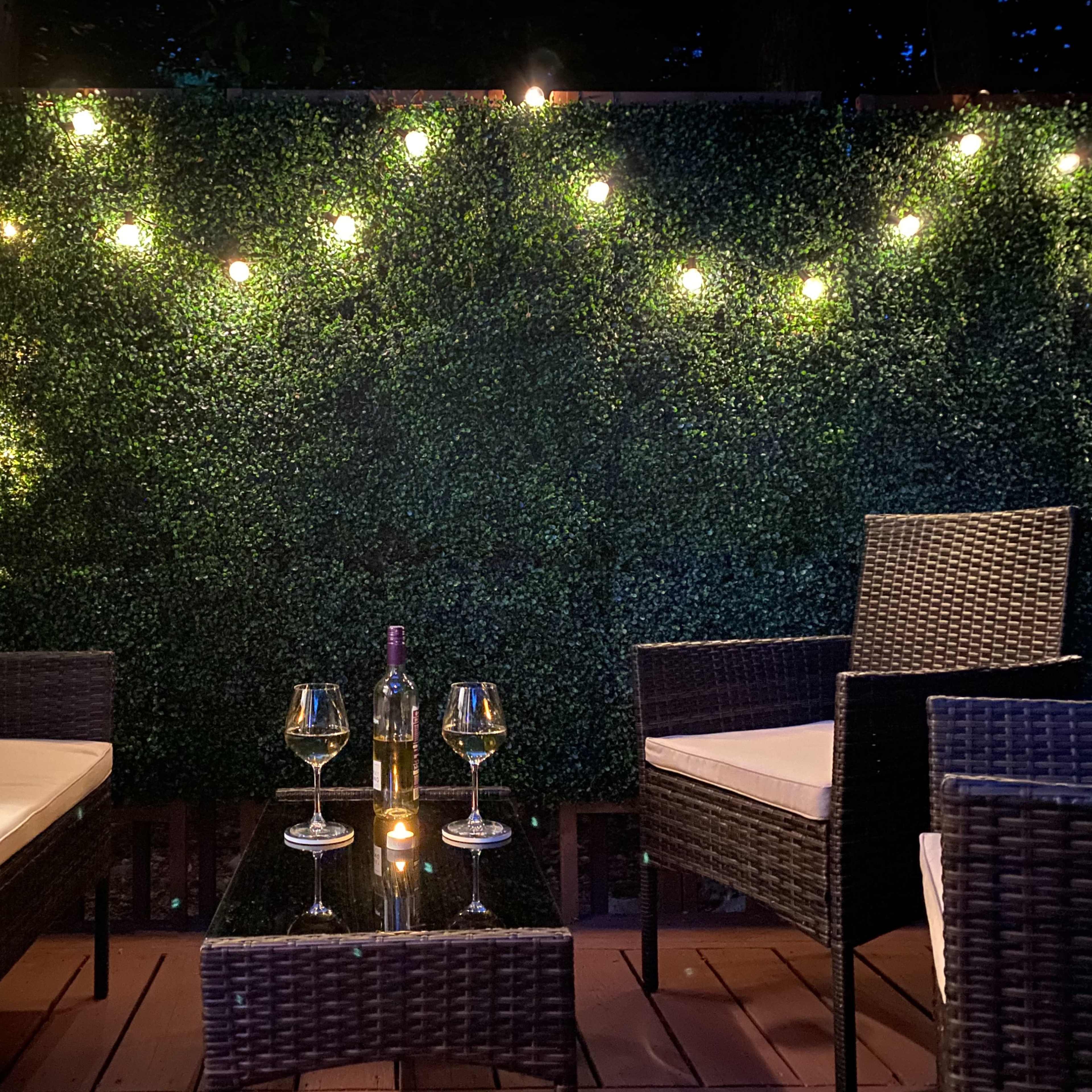A cozy outdoor seating area with two wicker chairs, a glass coffee table, a bottle of wine, and lit string lights against a green hedge backdrop.
